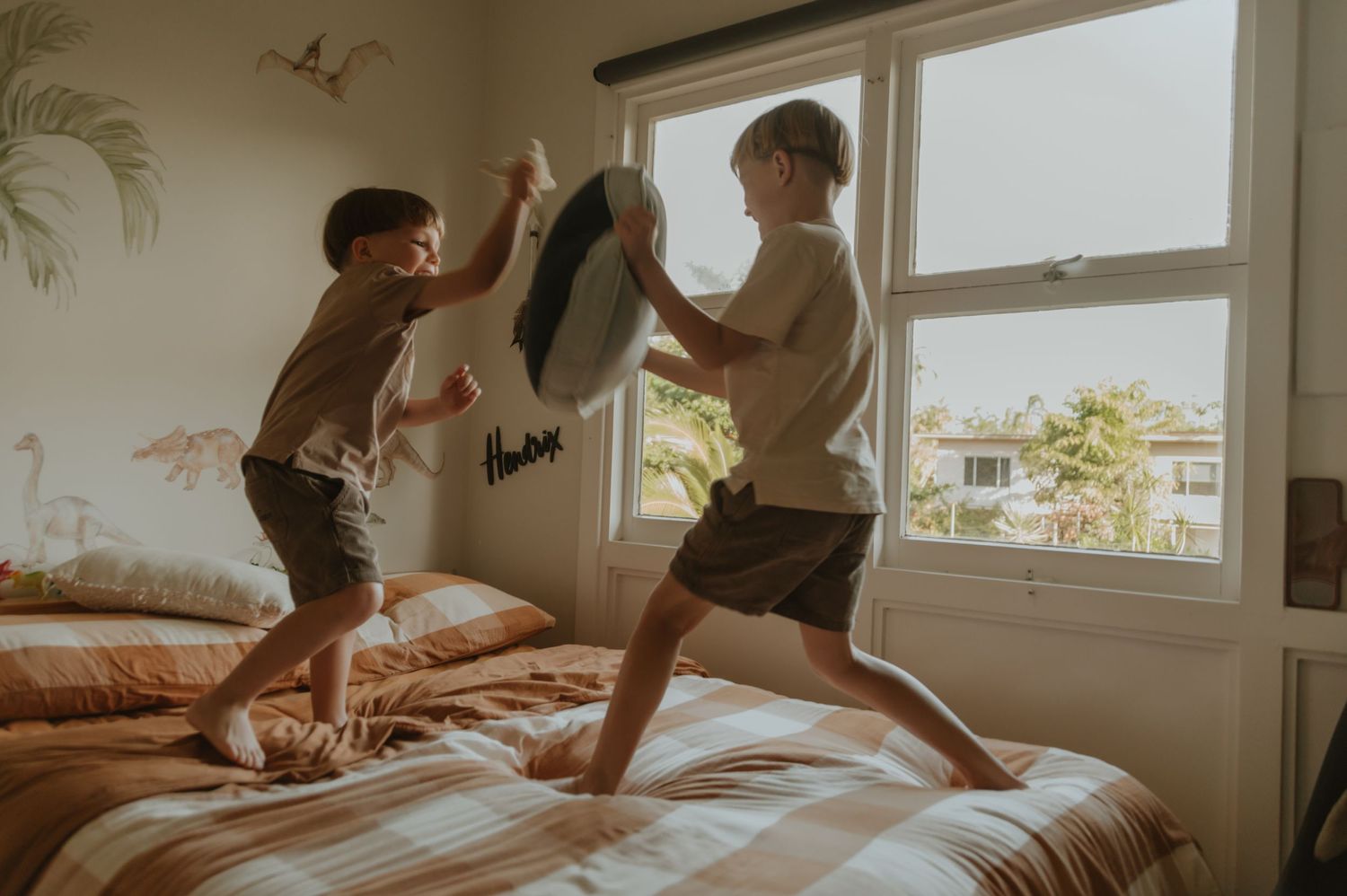 Two people jump playfully on a bed near a bright window in a sunlit bedroom.