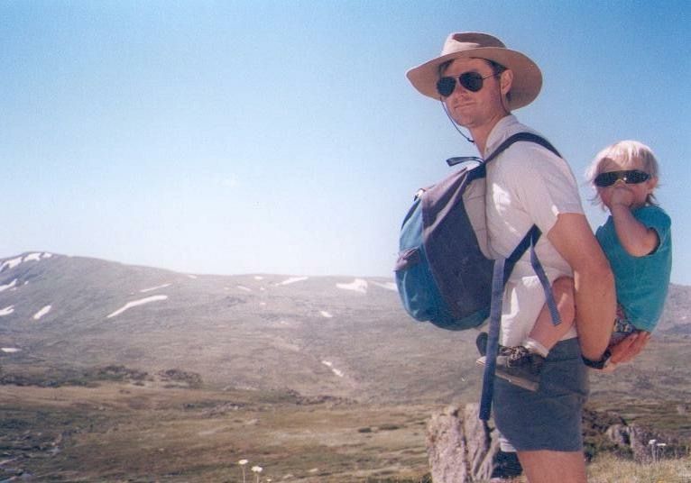 Two hikers wearing sunglasses and backpacks pause on a mountain trail with rugged terrain and blue sky in background.