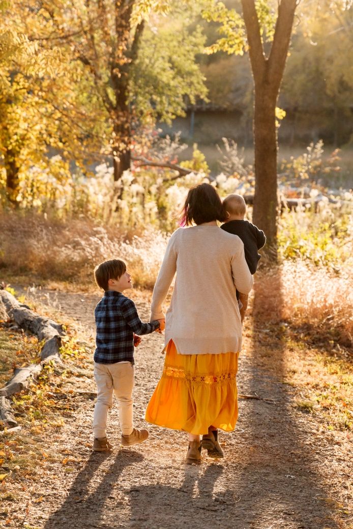 A mother holding a baby, with her child looking up at her in a sunlit autumn forest trail with figures casting long shadows in golden light.