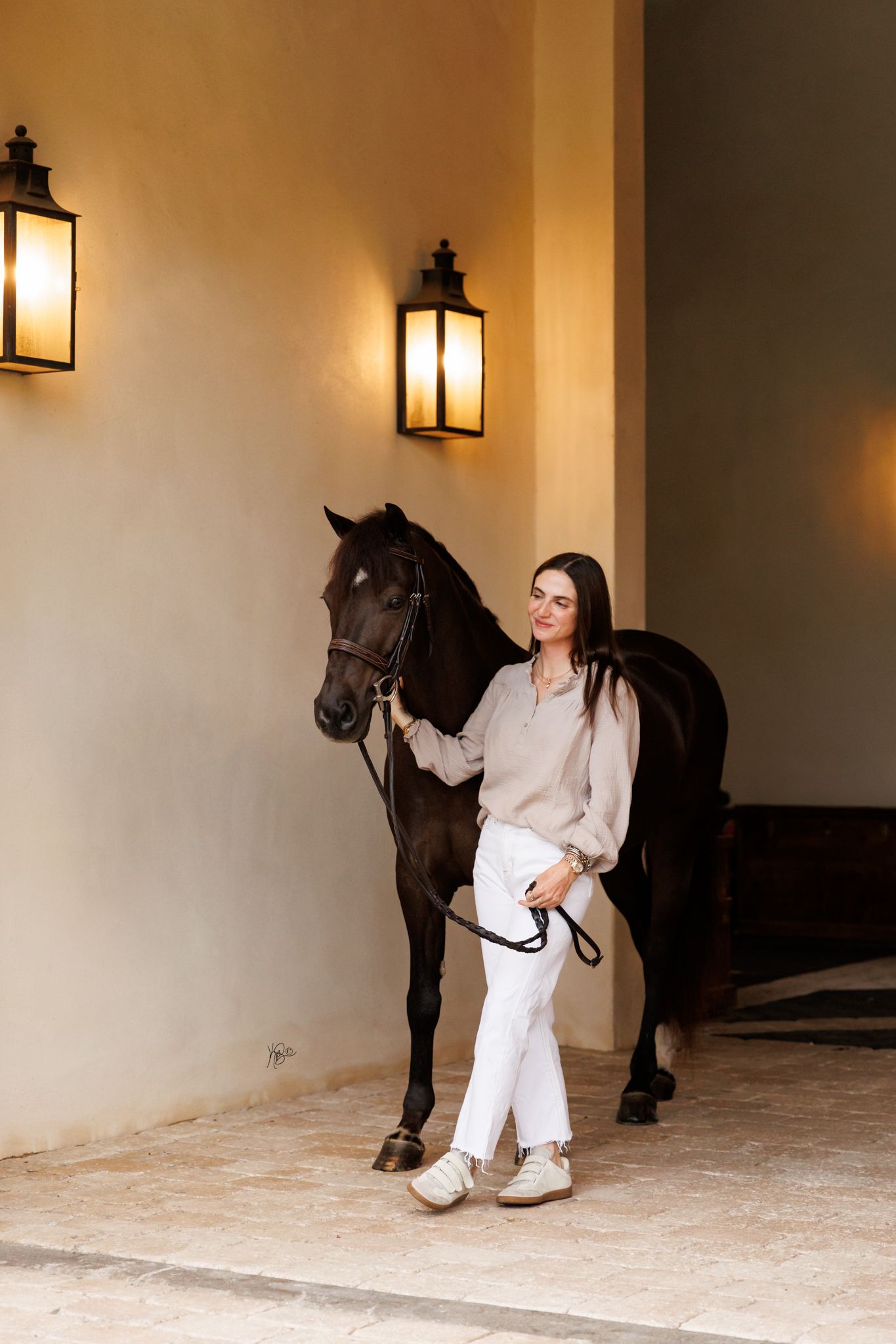 A woman in white pants and beige blouse poses with a dark horse through an elegant stable entrance with lanterns.