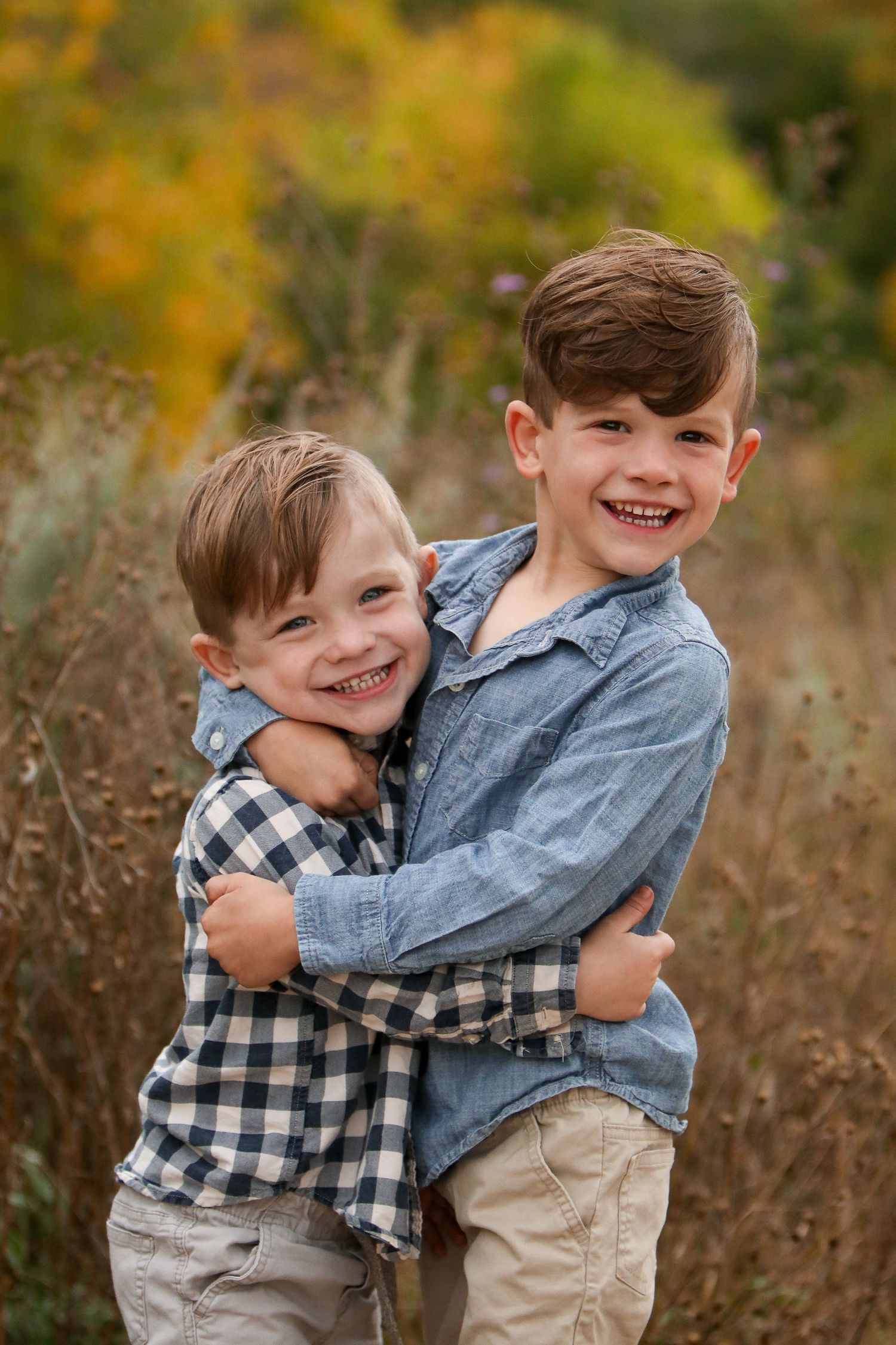 Two young brothers hugging and laughing in a field with fall colors in the background.