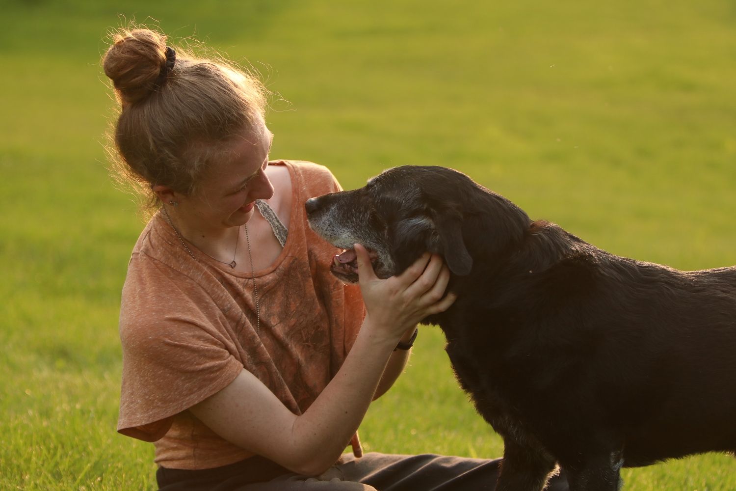 A girl in a peach colored shirt shares a tender moment with her black Labrador dog in a sunny green field.