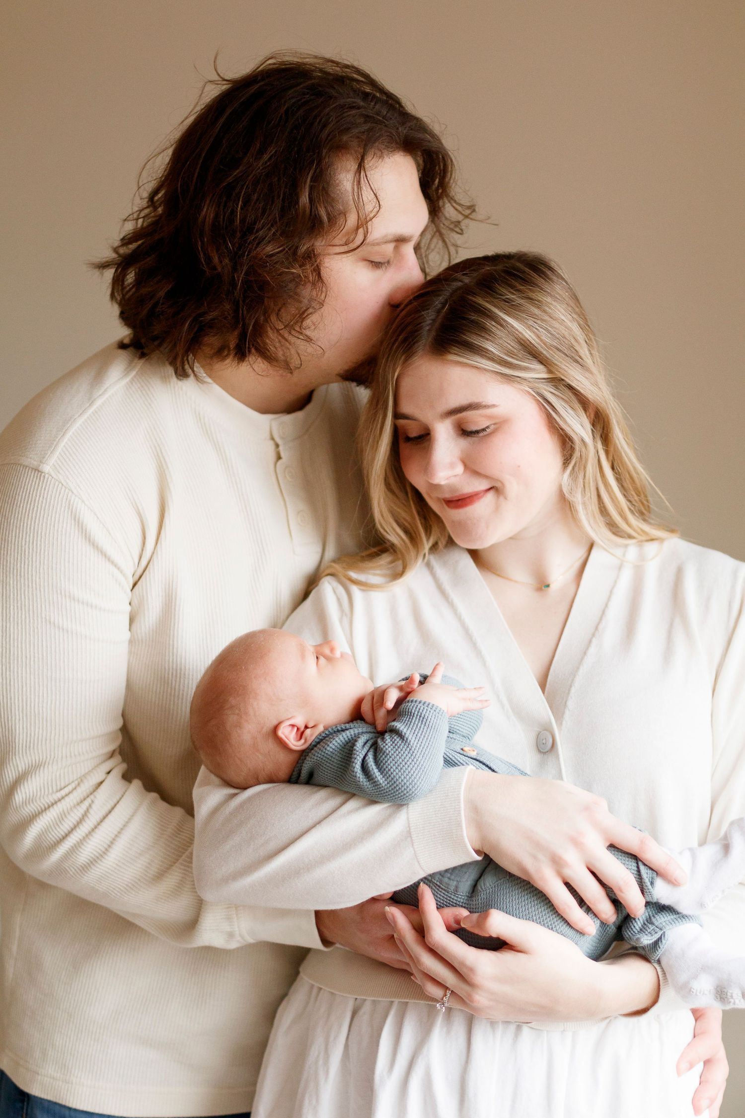 Intimate family portrait with newborn baby in soft white clothing against neutral backdrop.
