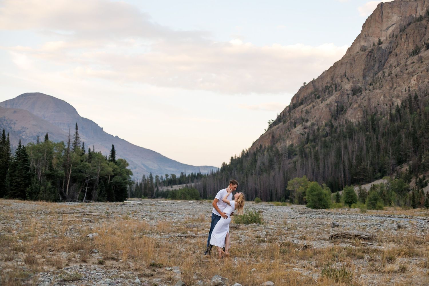 A young couple with the woman in a white dress embrace in front of a beautiful mountain setting