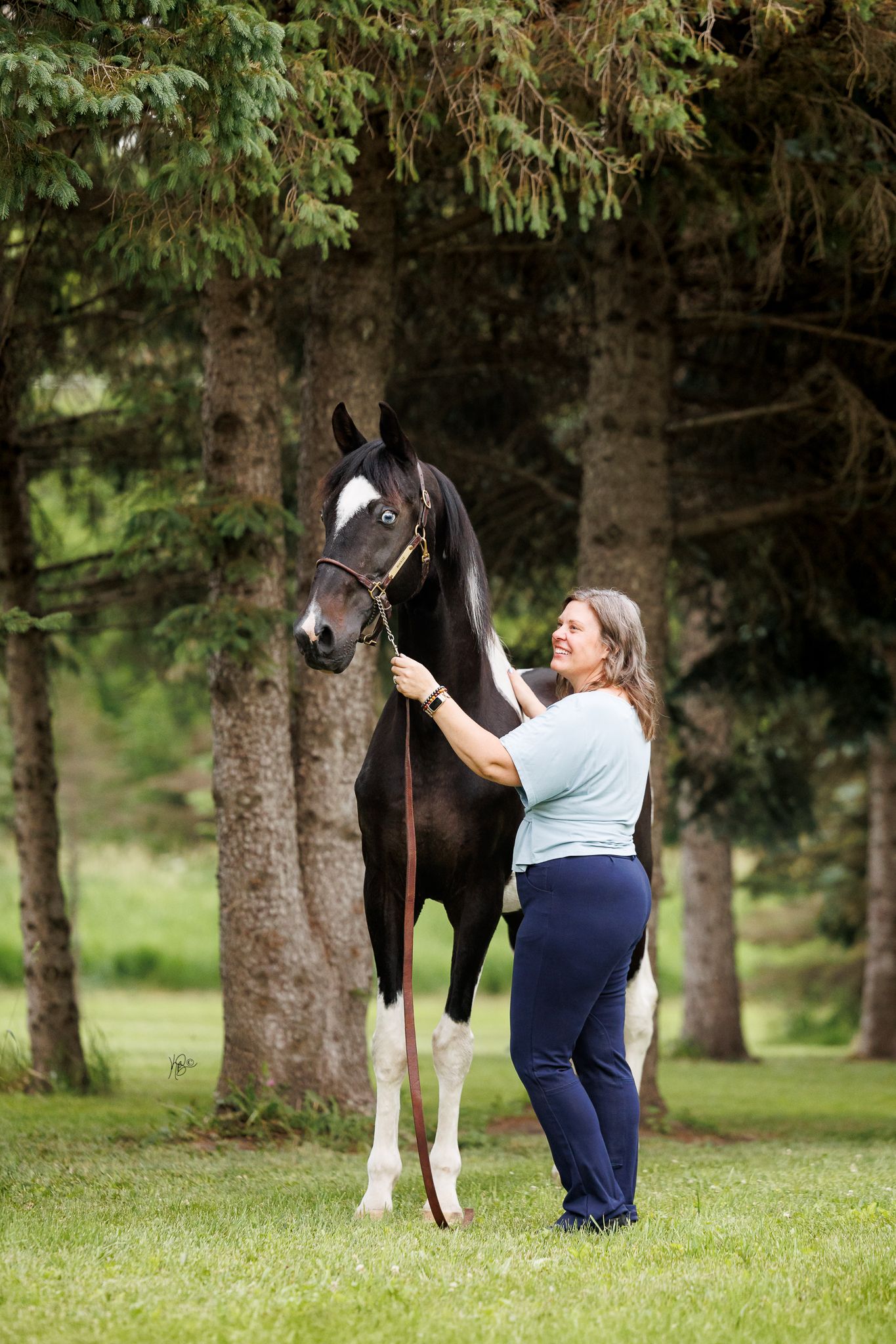 Woman smiling while holding the lead rope of a dark bay horse with a white blaze in a lush green park.