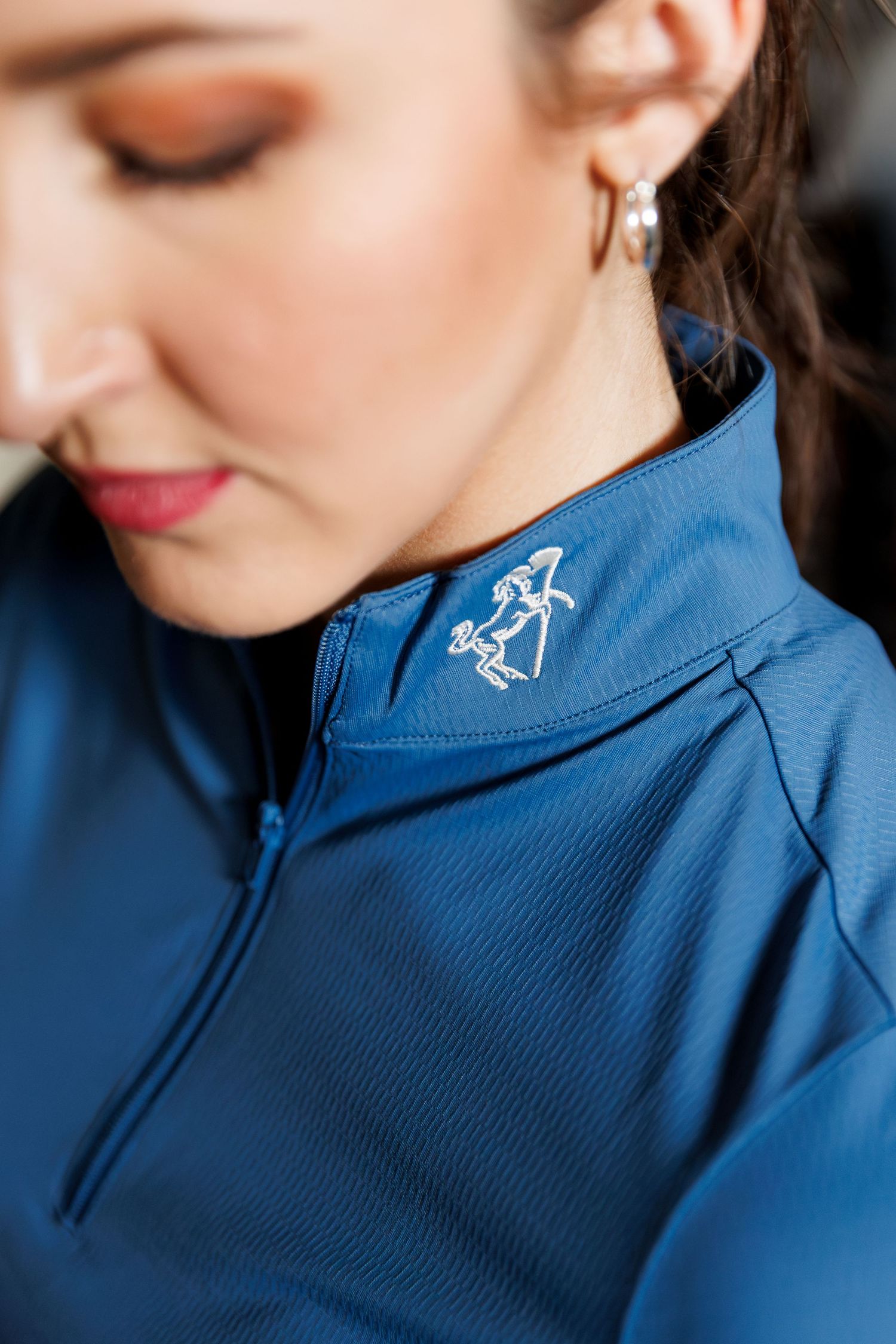 Woman wearing a blue quarter-zip riding shirt with an embroidered logo, paired with silver hoop earrings.