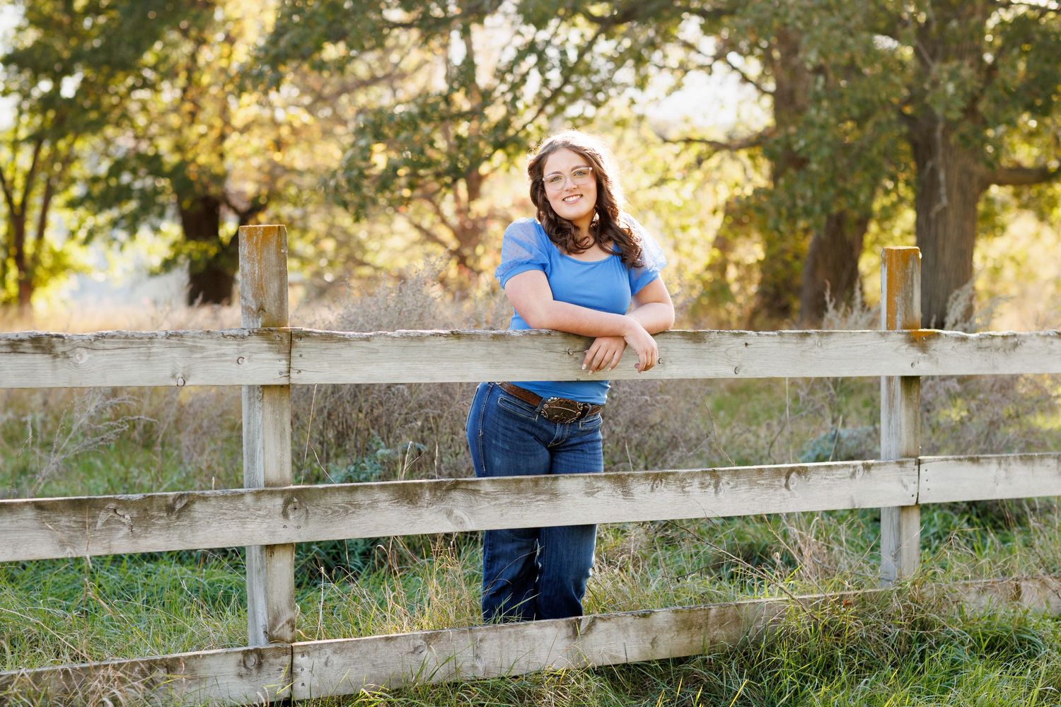 A rustic wooden fence stretches across a grassy field at sunset while a young woman poses for senior portraits.