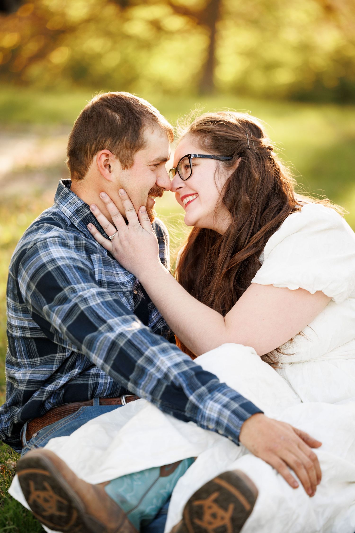 A couple sitting on grass, smiling and touching foreheads in a sunny park, woman wearing glasses and white dress.