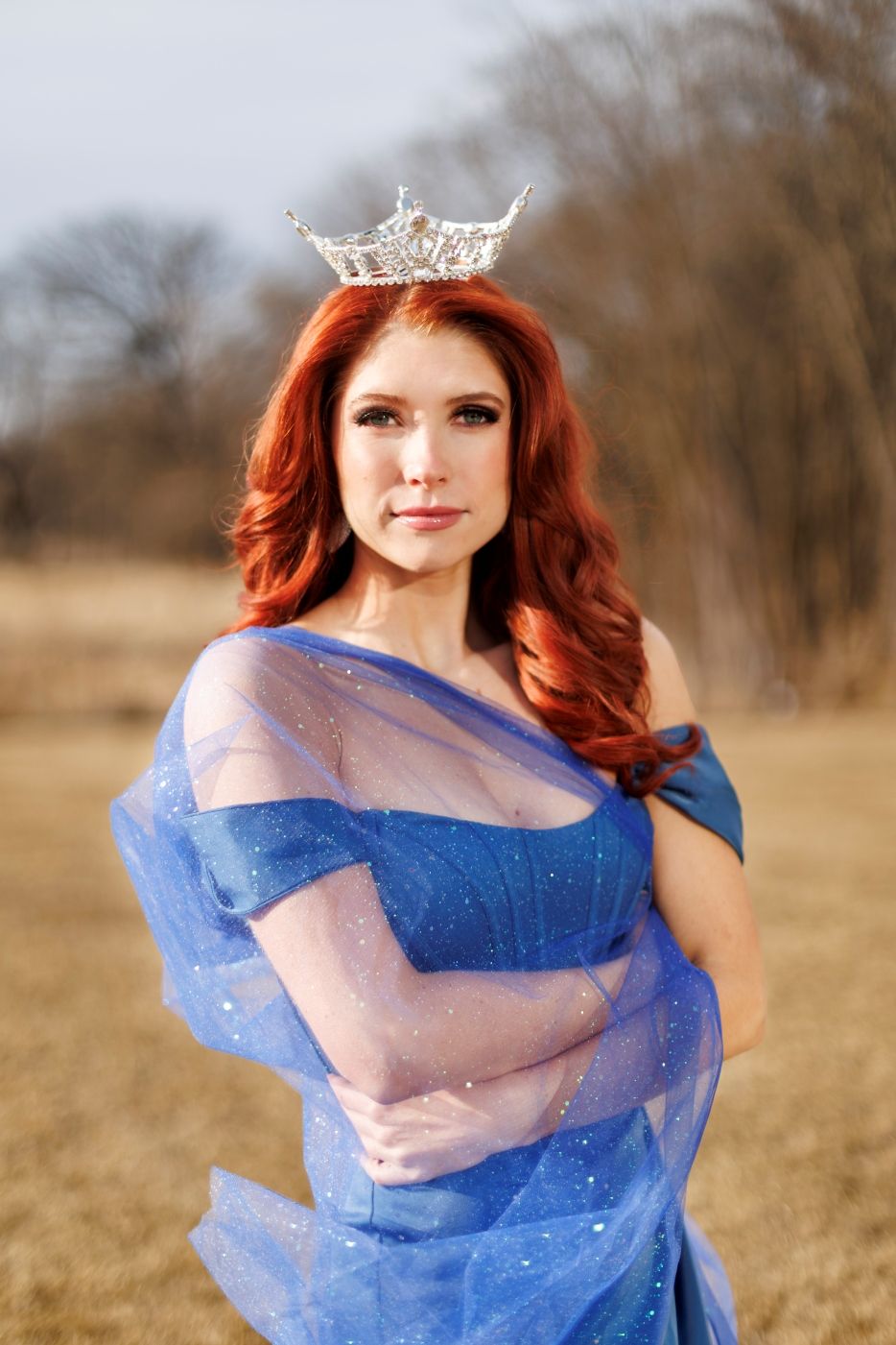 Young woman in flowing blue sparkly dress and crown poses in an open field during golden hour.