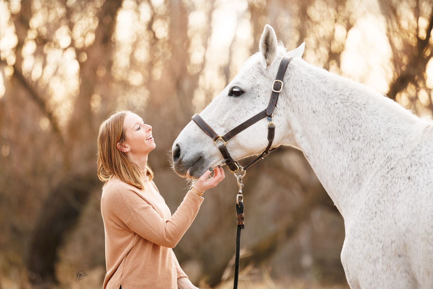 A woman in a beige sweater shares a tender moment with a white horse during a golden sunset.