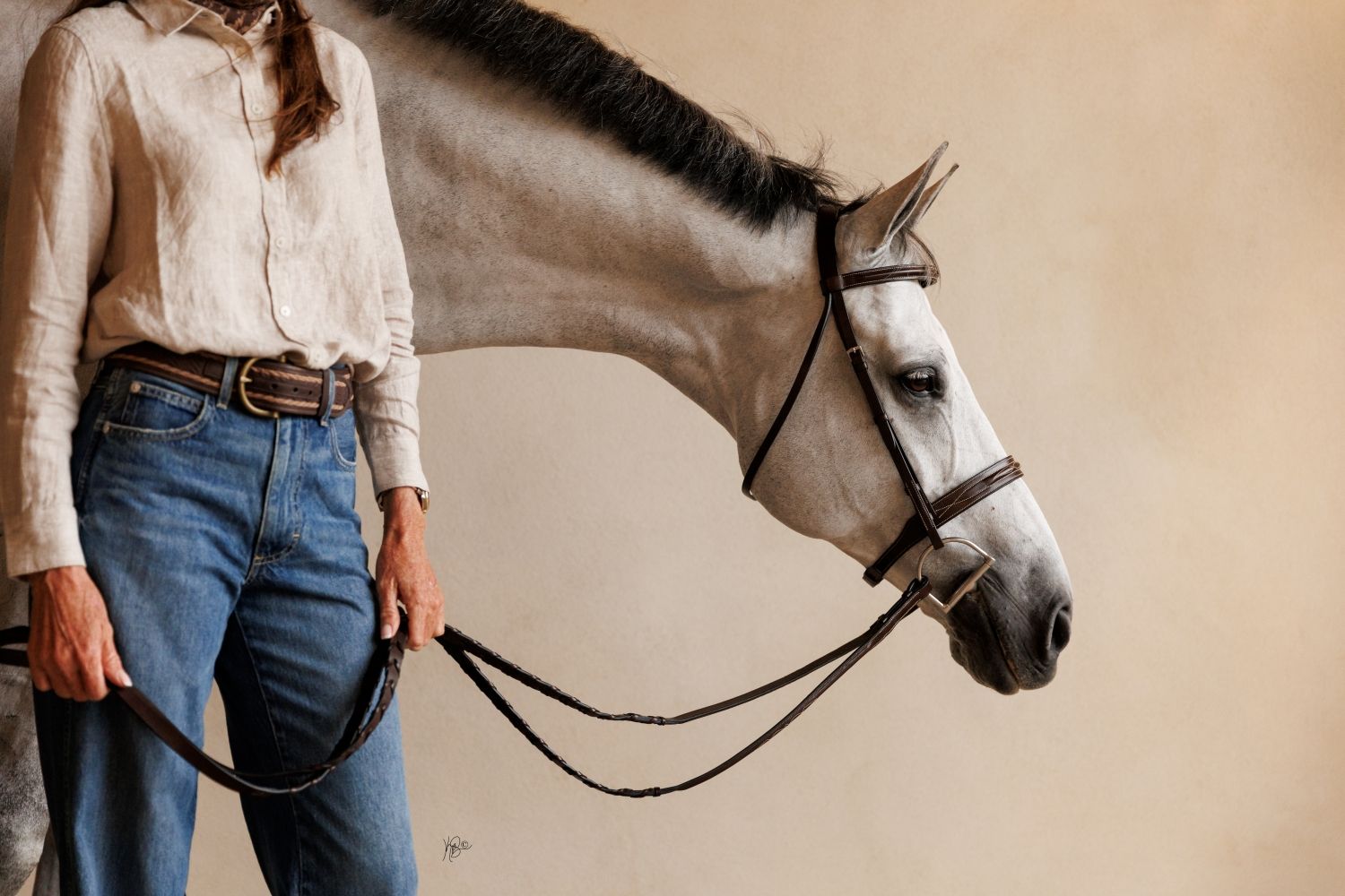 A chic, editorial portrait of a woman standing beside a grey horse with a bridle against a neutral background.