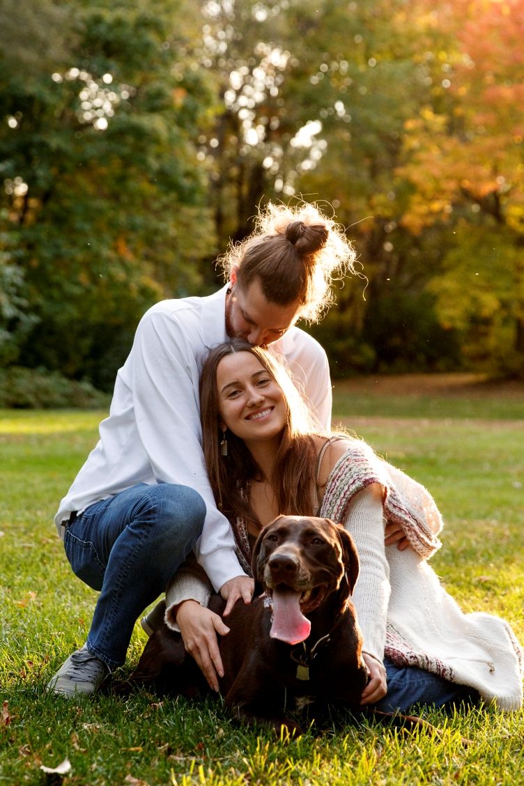 Happy couple embracing sitting on grass with their chocolate lab dog during golden hour.