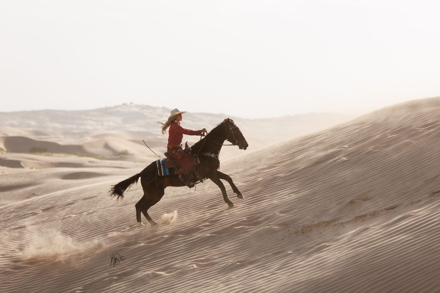 Horse and rider gallop dramatically across desert sand dunes at sunset.