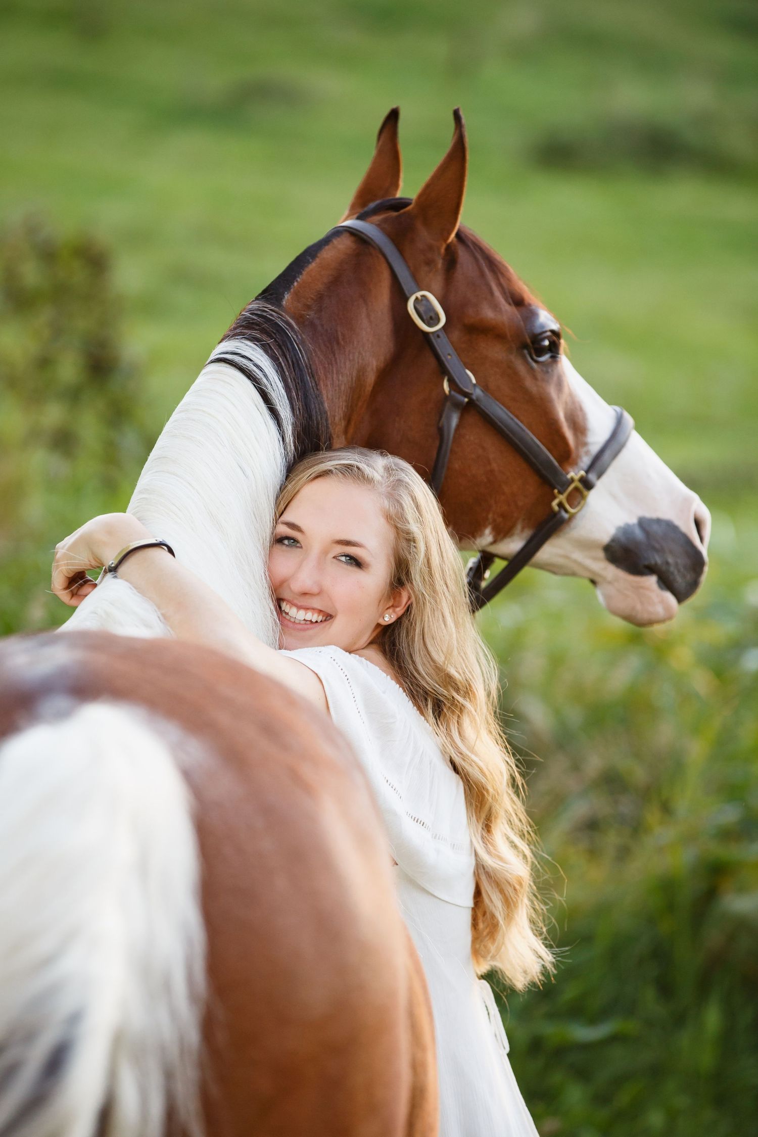 Beautiful brown and white pinto horse standing in green field being embraced by a woman in a white dress