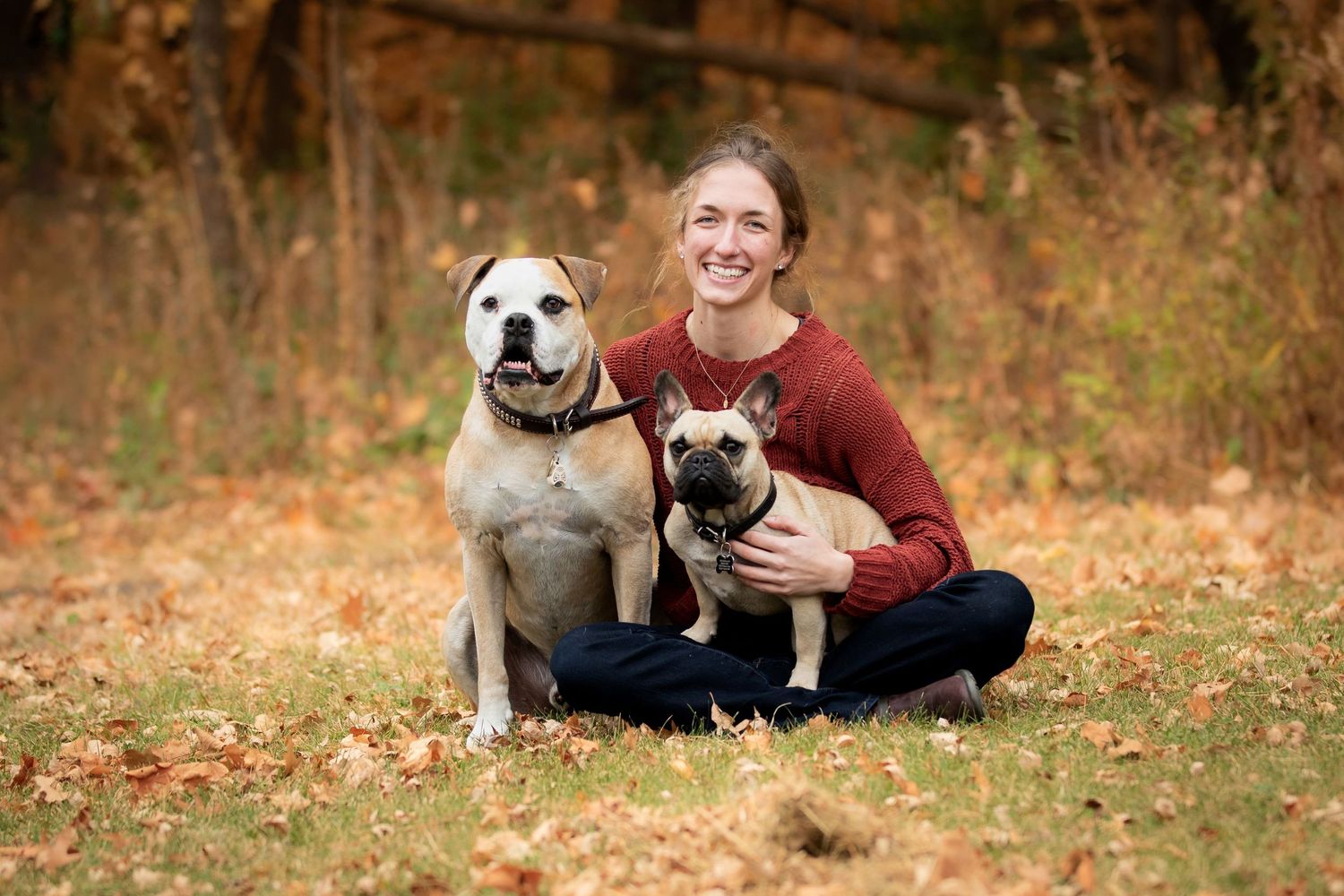 Me, a young woman, in a burgundy sweater sitting with two dogs, an american bulldog a smaller French Bulldog, on autumn ground.
