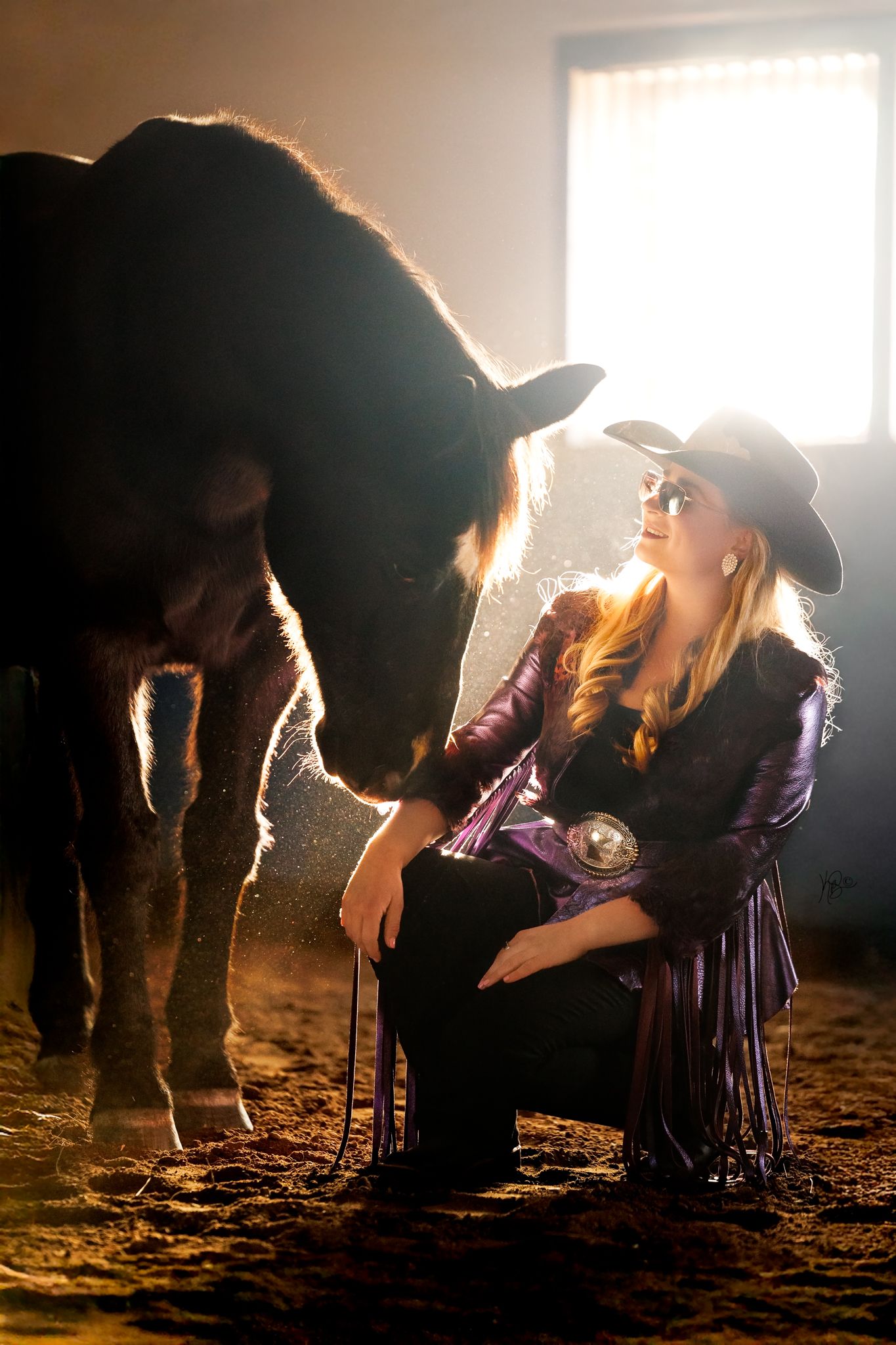 A horse and rider share an intimate moment in a sunlit barn with dramatic backlighting.