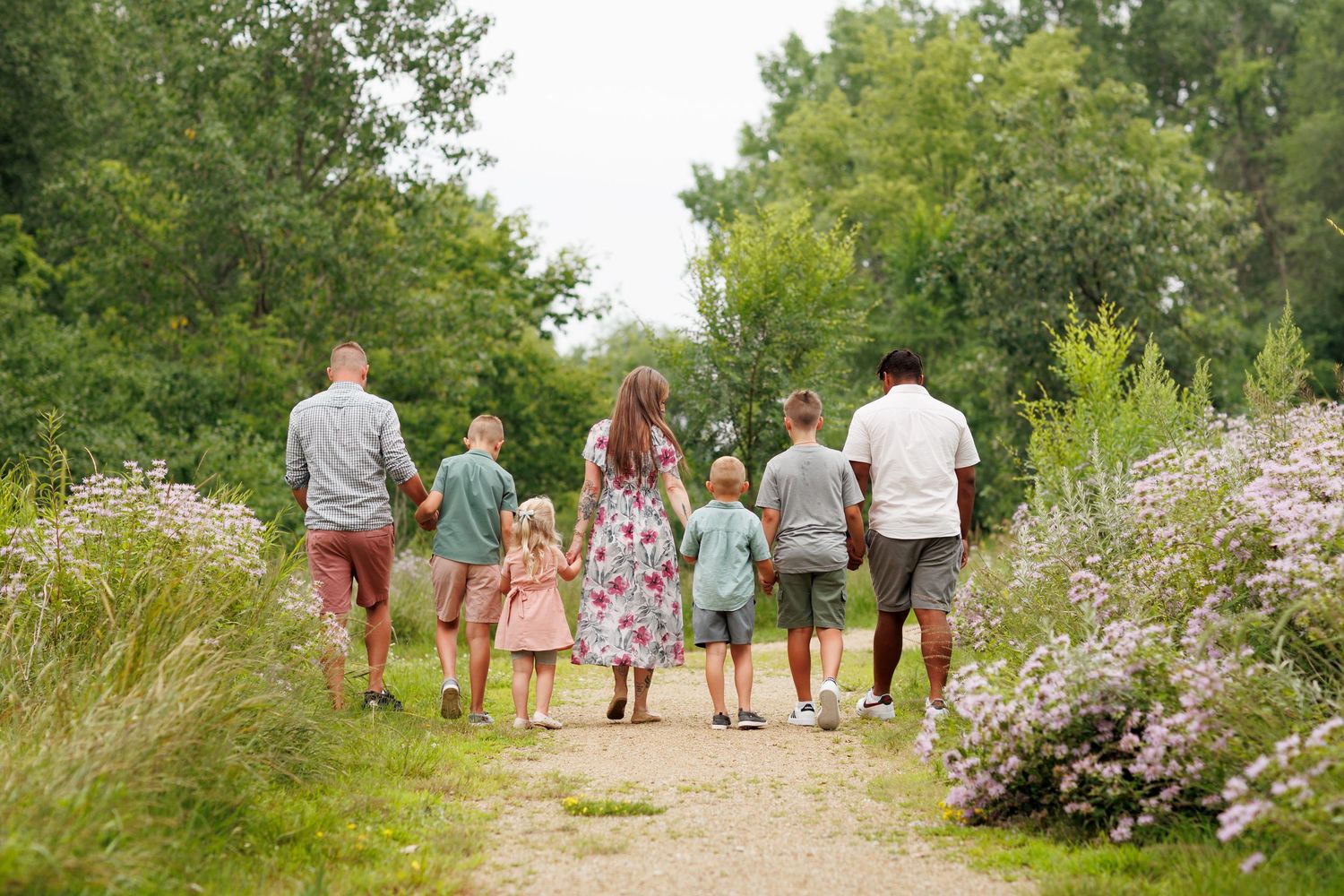 A family walking together on a scenic nature trail surrounded by blooming wildflowers and green trees on a summer day.