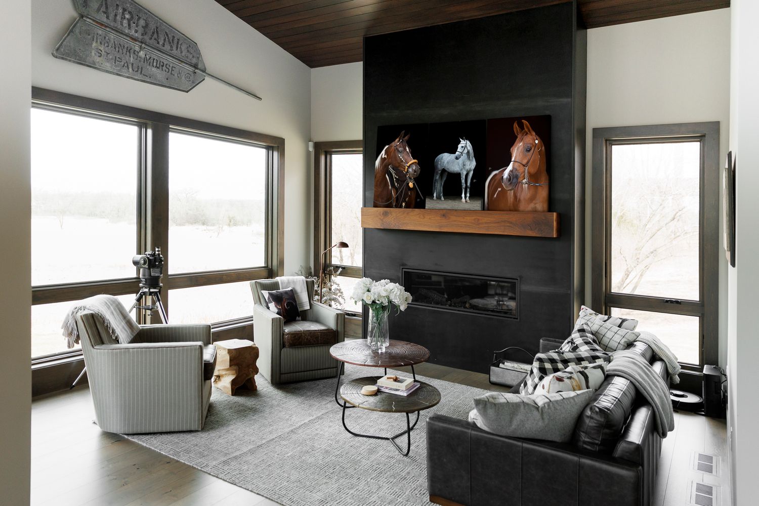 Modern living room interior with dark walls, wood ceiling, and large windows showcasing three large prints of horse portraits.