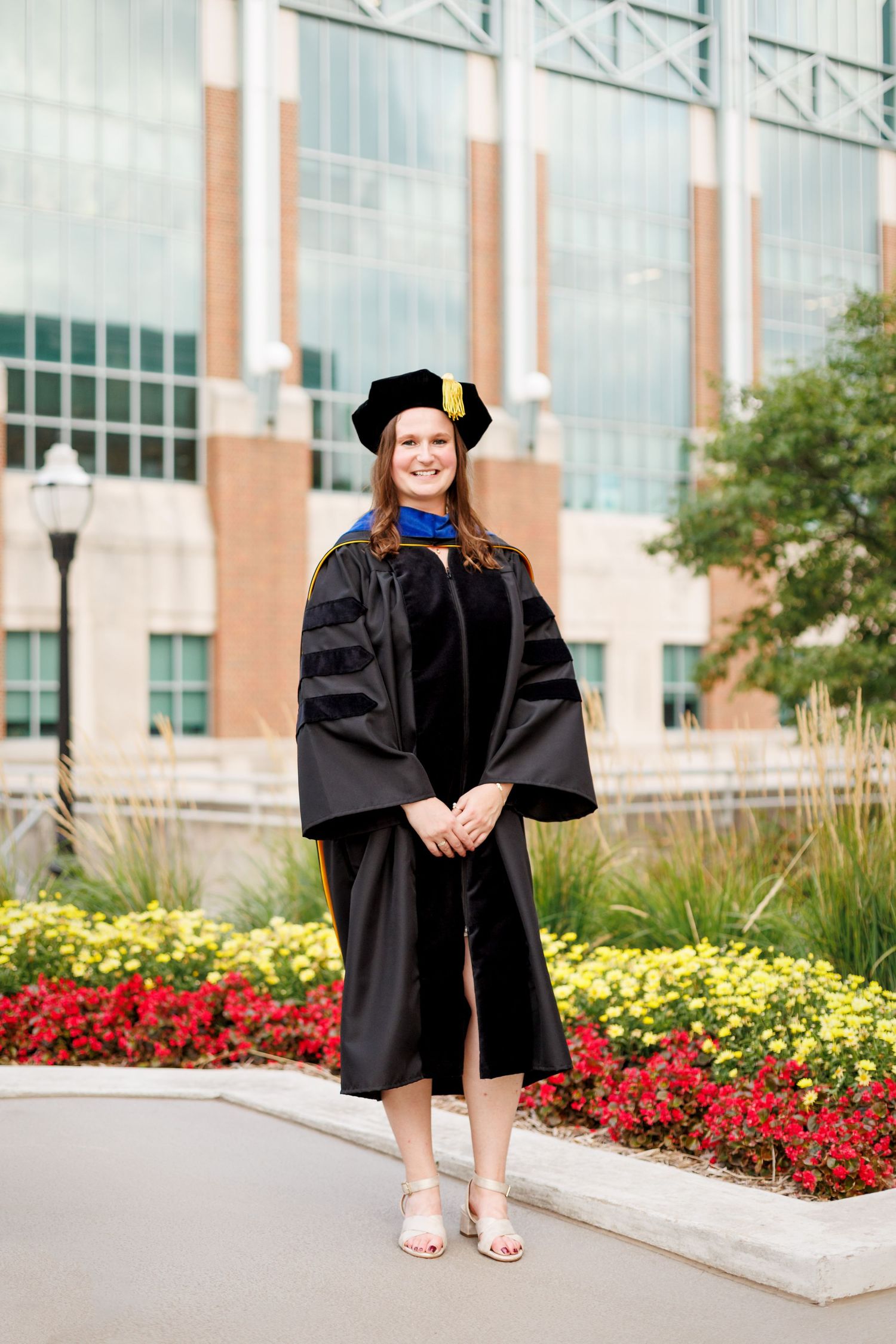 Graduate in black cap and gown poses for photos near colorful flower beds and modern campus buildings.