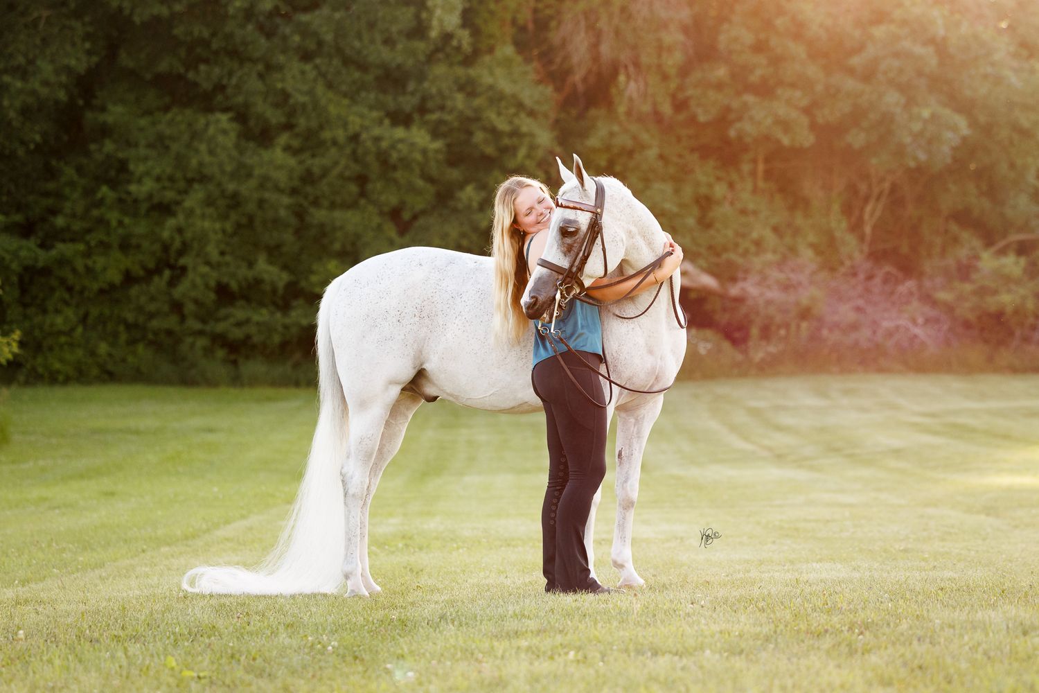 A blonde woman stands beside a white horse on a lush green lawn with trees in the background.