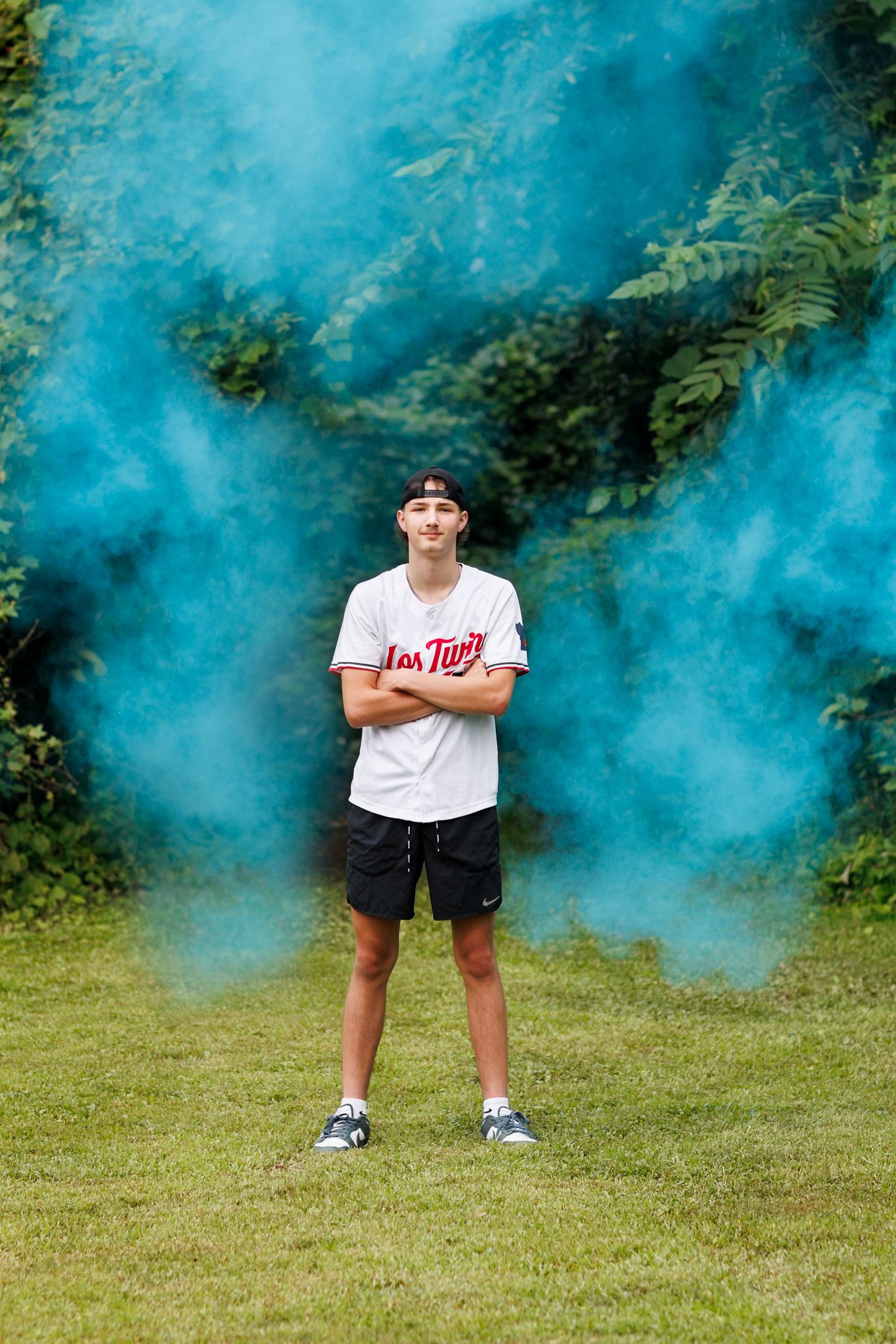 High school senior guy standing in front of bright blue smoke in garden setting.