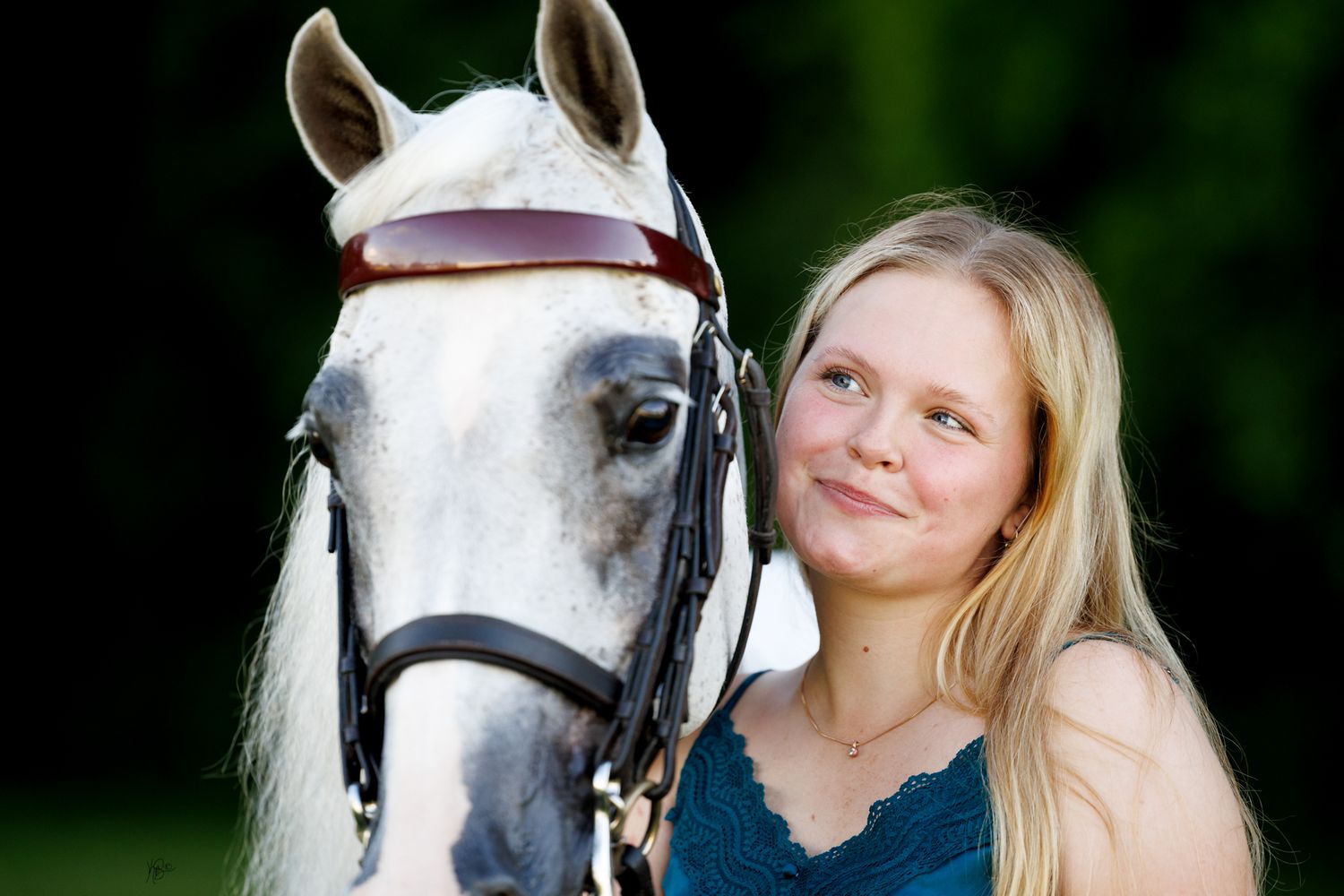 Young blonde girl smiling beside a white horse wearing a bridle, outdoors with green background.