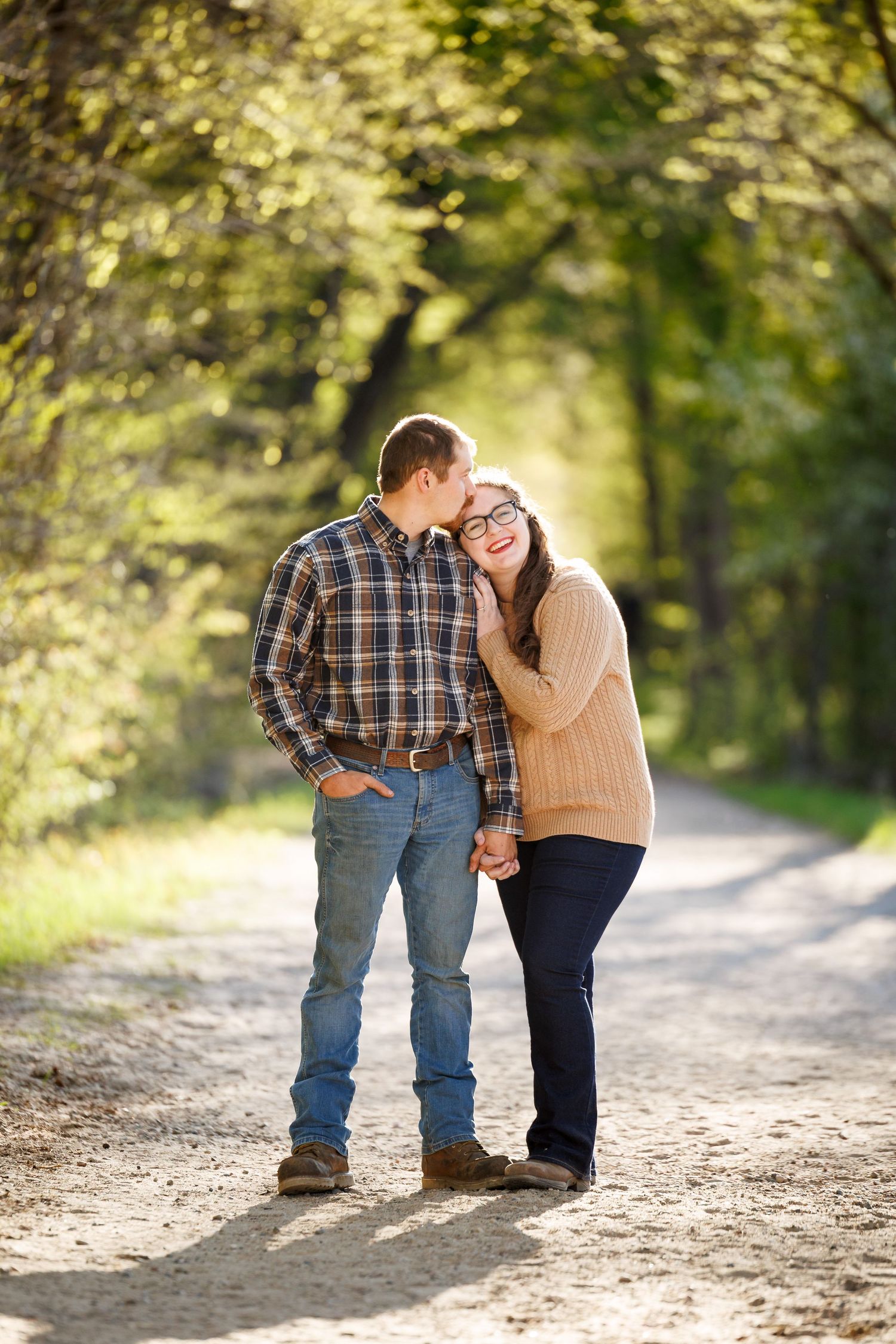 A couple holds hands and shares a tender moment on a sunlit dirt path surrounded by lush green trees.