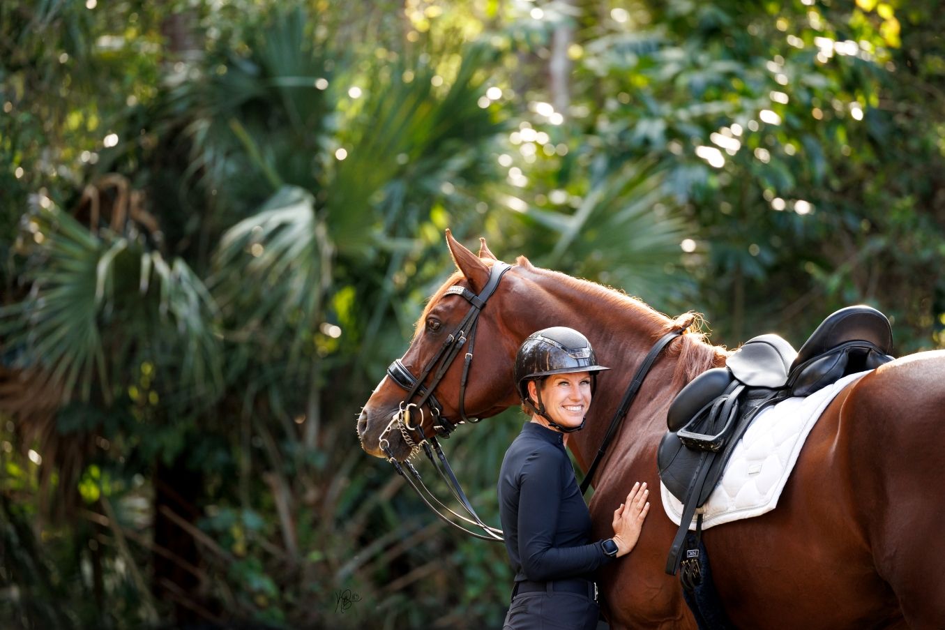 A smiling equestrian in a black helmet embraces a chestnut horse amid lush green tropical foliage.