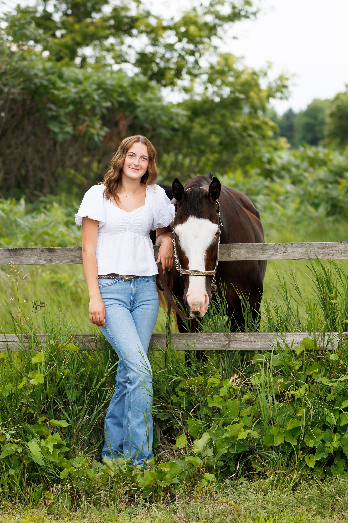 Senior equestrian with her brown horse stand behind a wooden fence in a lush green summer pasture.