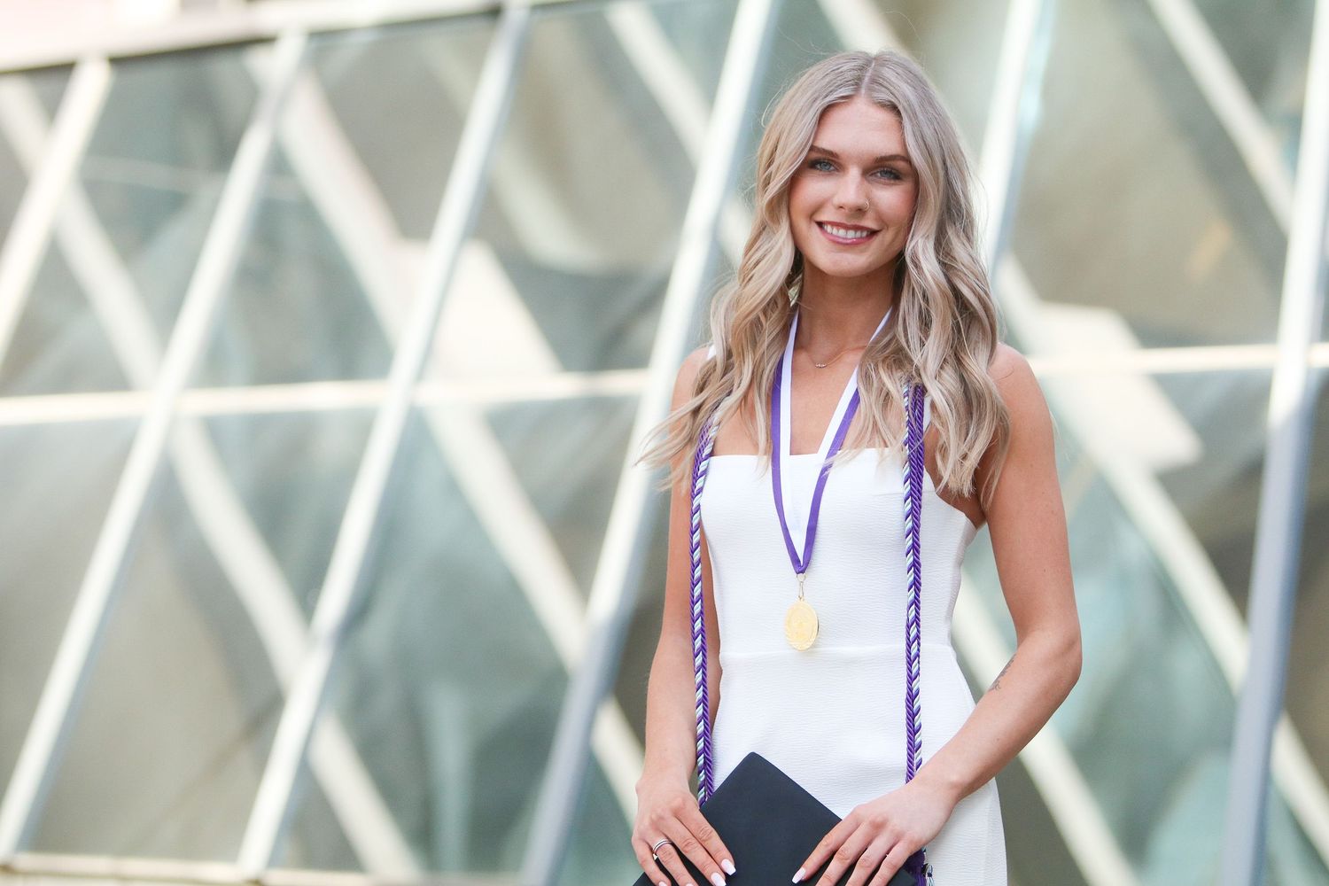 College graduate in white dress holding diploma with geometric architecture in background.