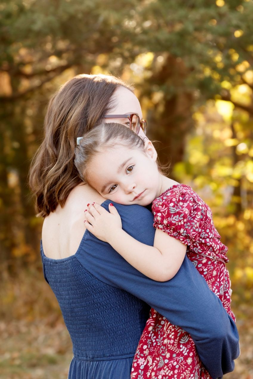 Tender moment of a young girl on her mom's shoulder captured outdoors with warm autumn colors in the background.
