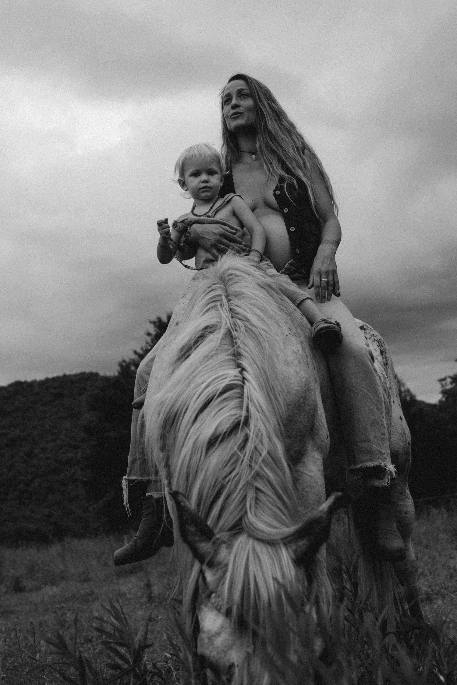 Black and white photo of a woman and toddler riding a light-colored horse in a field under cloudy skies. Maternity photographer Javea Spain