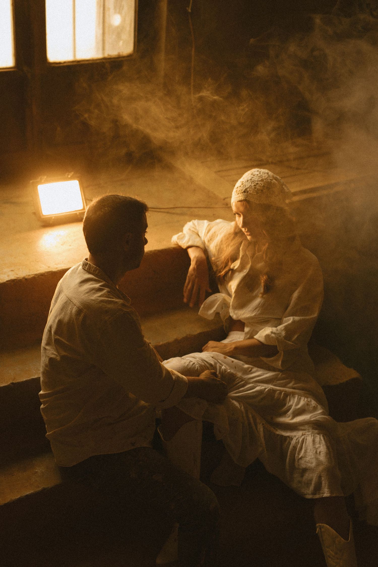 Two people in white clothing sit together in a misty, warmly lit rustic room, engaged in an intimate conversation. Couple photographer Javea