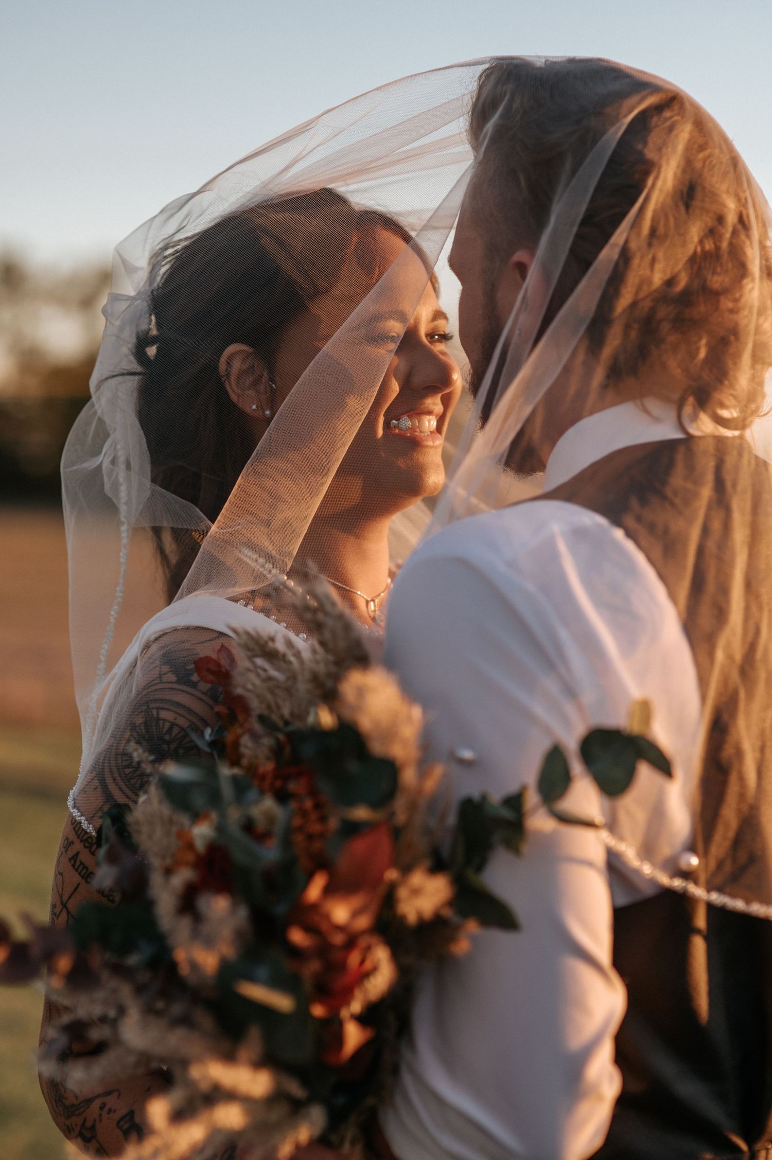 Two newlyweds share an intimate moment under flowing veils during golden hour sunset wedding photography session.