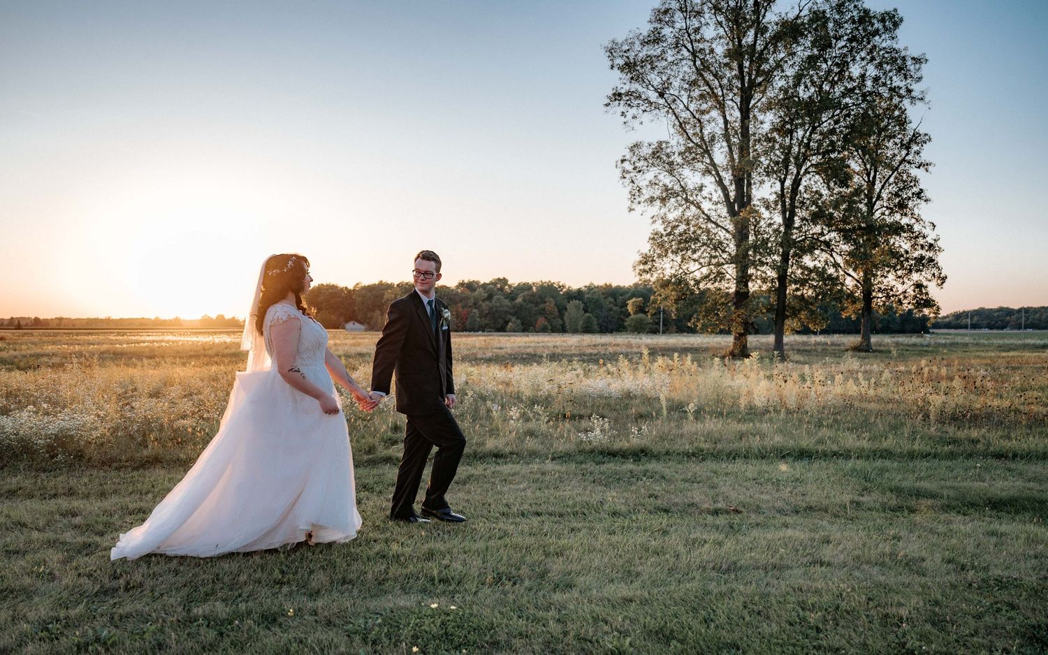 A wedding couple walks through a golden field at sunset while their photographer captures romantic moments.