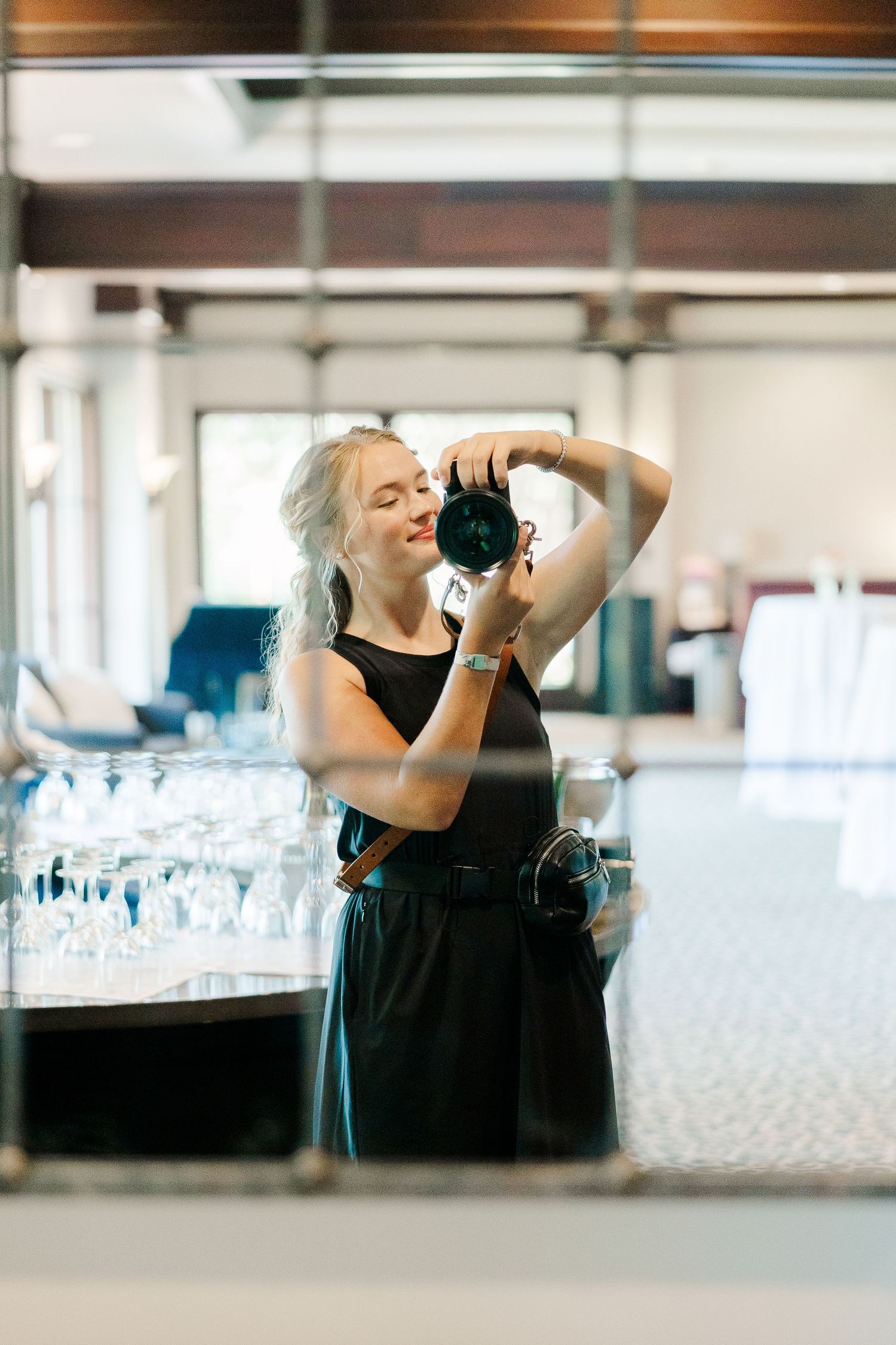 Blonde woman in black dress photographing with DSLR camera near elegant glassware display in modern venue.