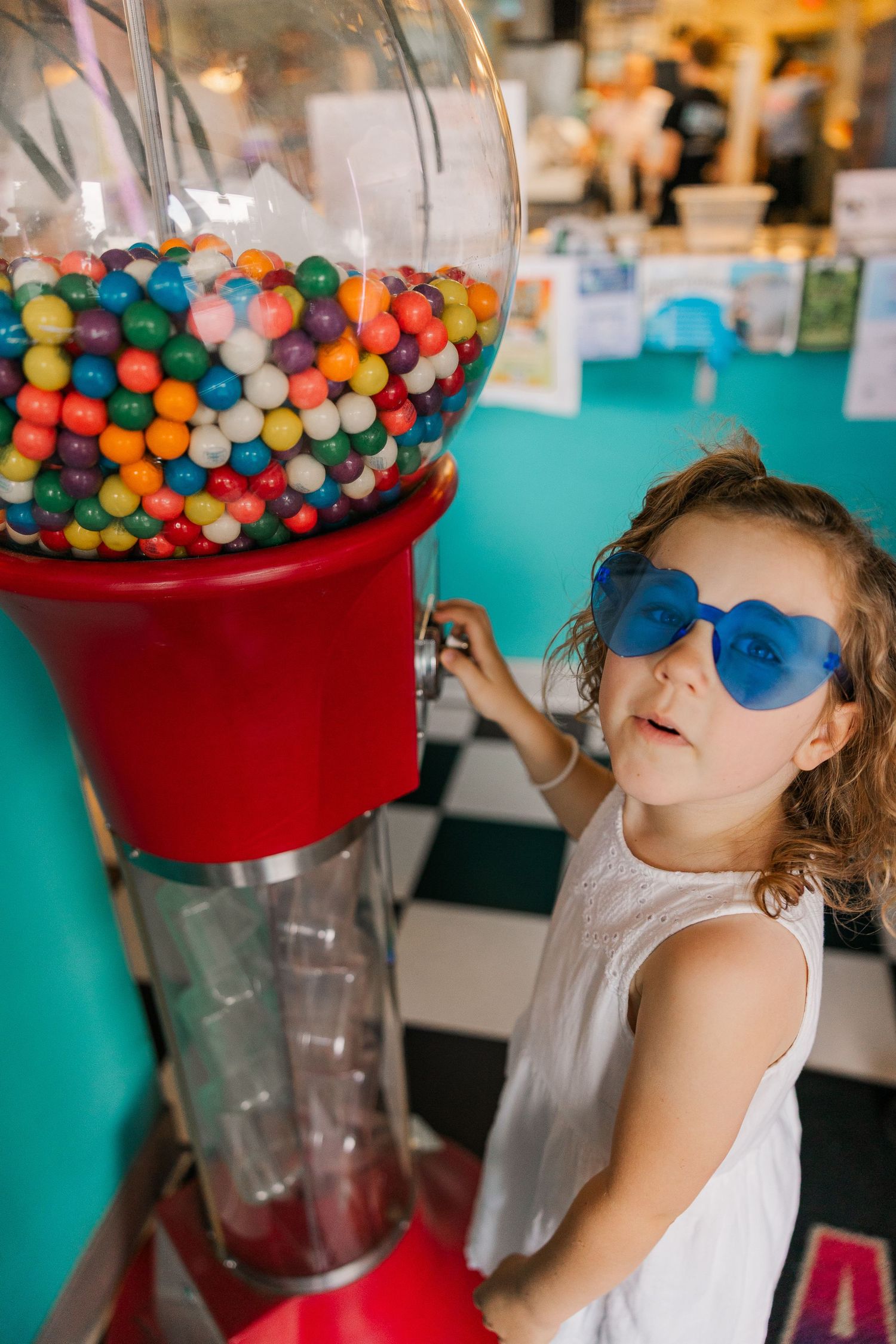 Young girl wearing blue heart sunglasses reaching for a colorful gumball machine in a retro ice cream shop.