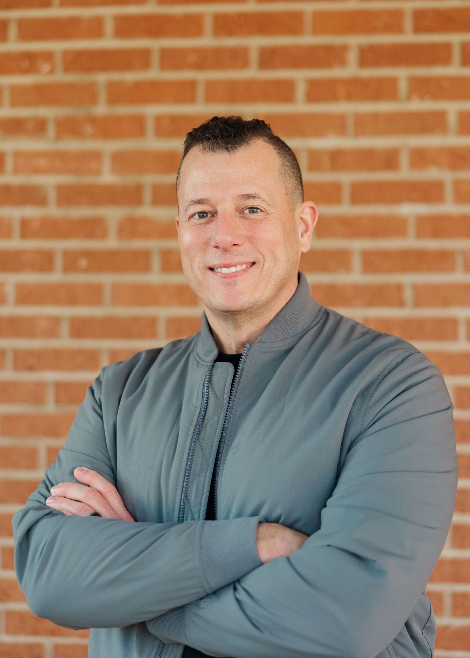 Man in gray zip-up jacket smiling with arms crossed against a red brick wall background.