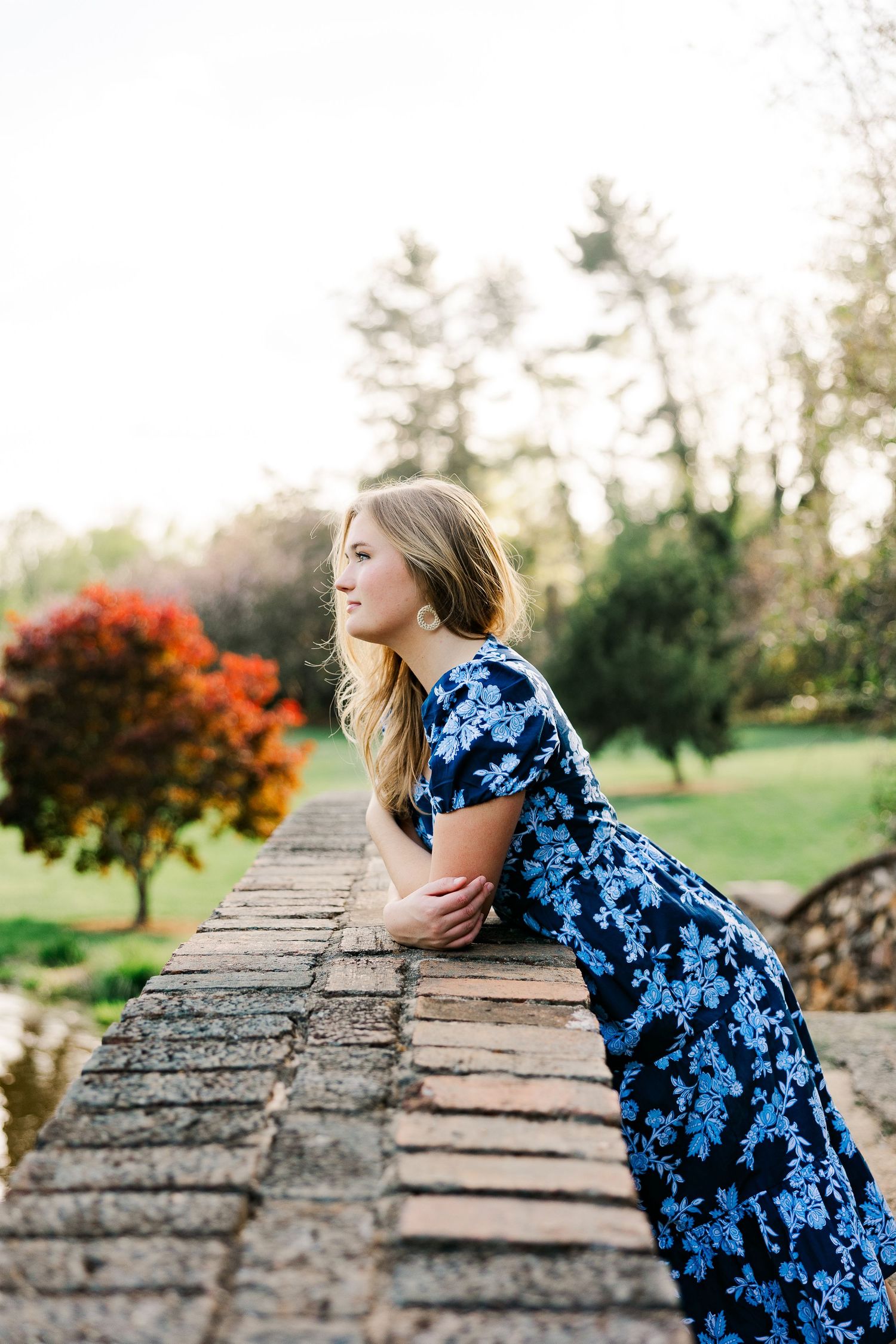 Young woman in blue floral dress leaning on stone wall in lush garden, smiling softly in natural light.