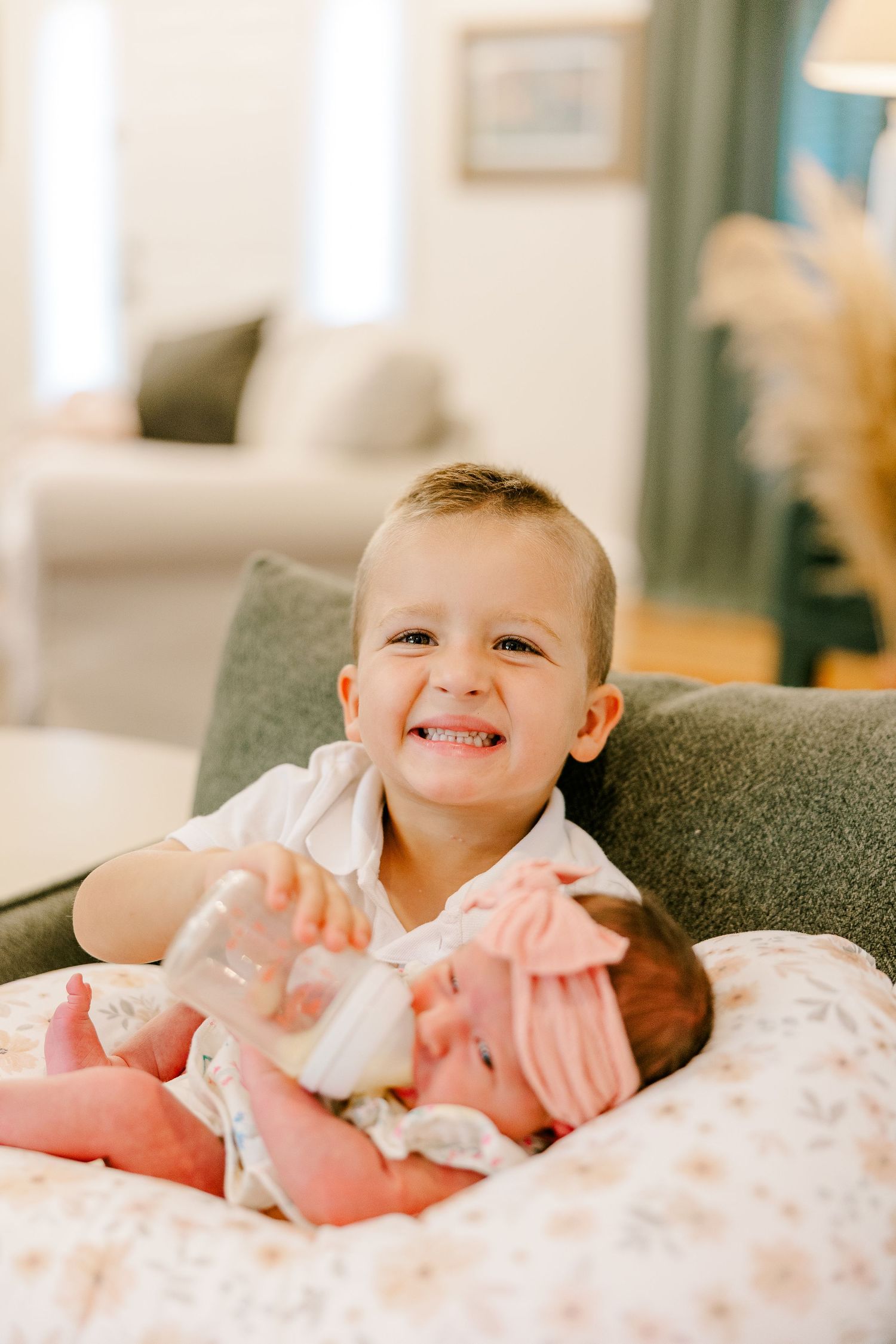Smiling toddler boy holding a baby bottle while cradling a newborn sibling on a nursing pillow indoors.