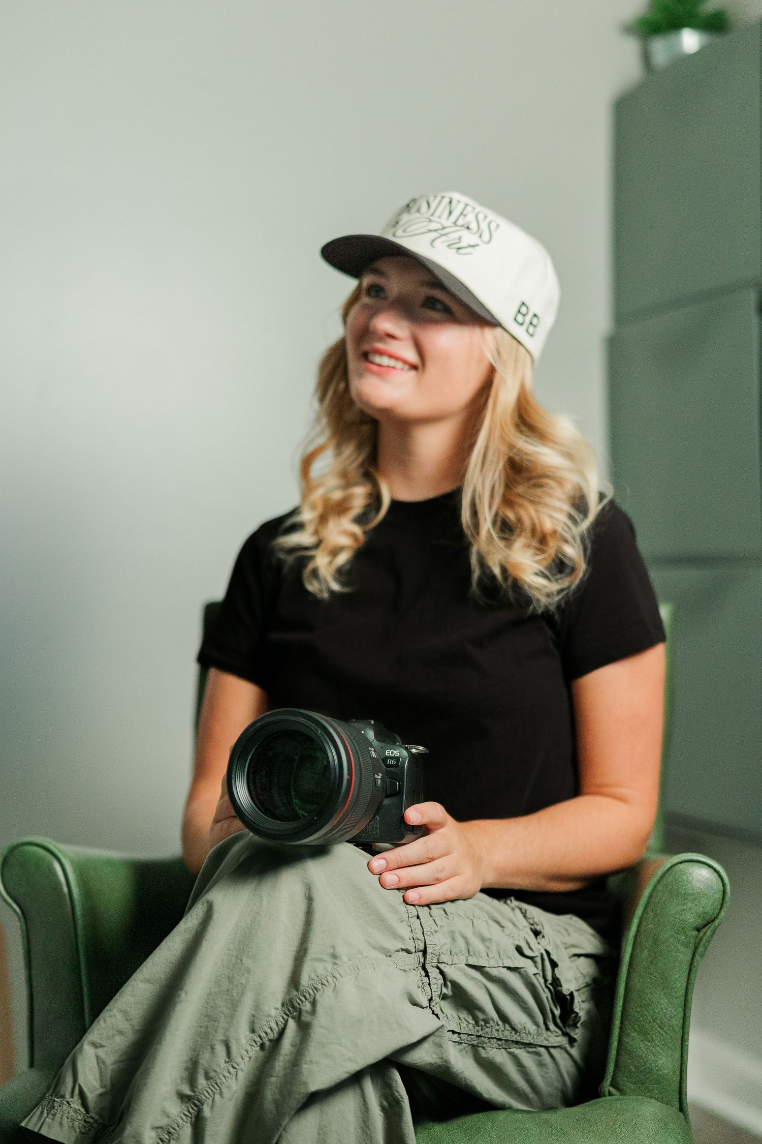 Smiling blonde woman in black tee and white cap holds a camera while seated in a green leather chair.