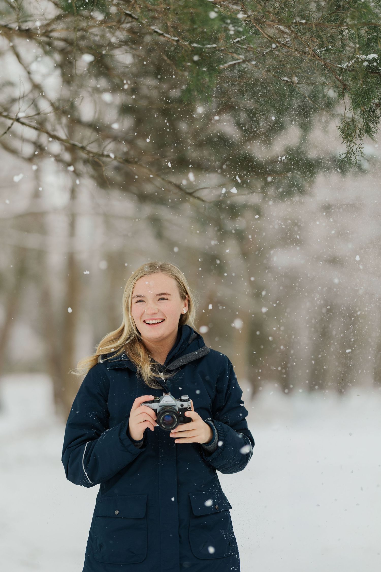 A photographer wearing a navy winter coat laughs joyfully while taking pictures in a snowy forest setting.