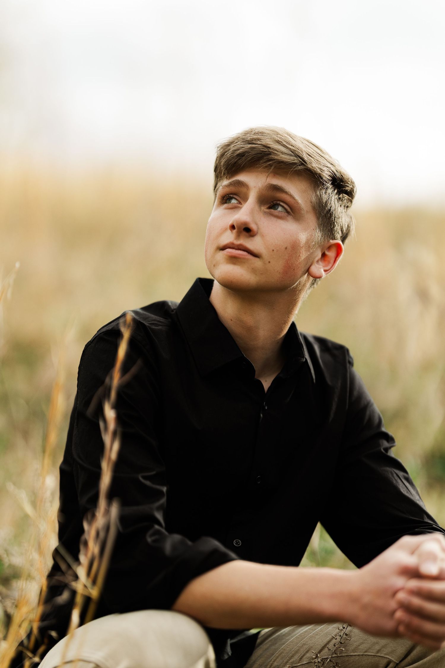 Young man in black shirt sitting in golden grass field, gazing upward thoughtfully in outdoor portrait.