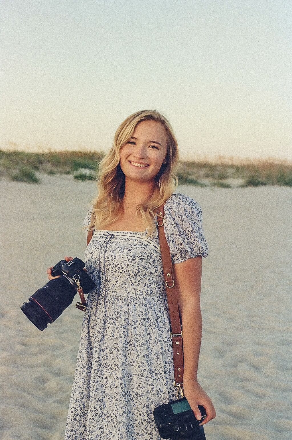 Smiling blonde woman in a floral sundress holds a professional camera on a sandy beach with dunes at sunset.
