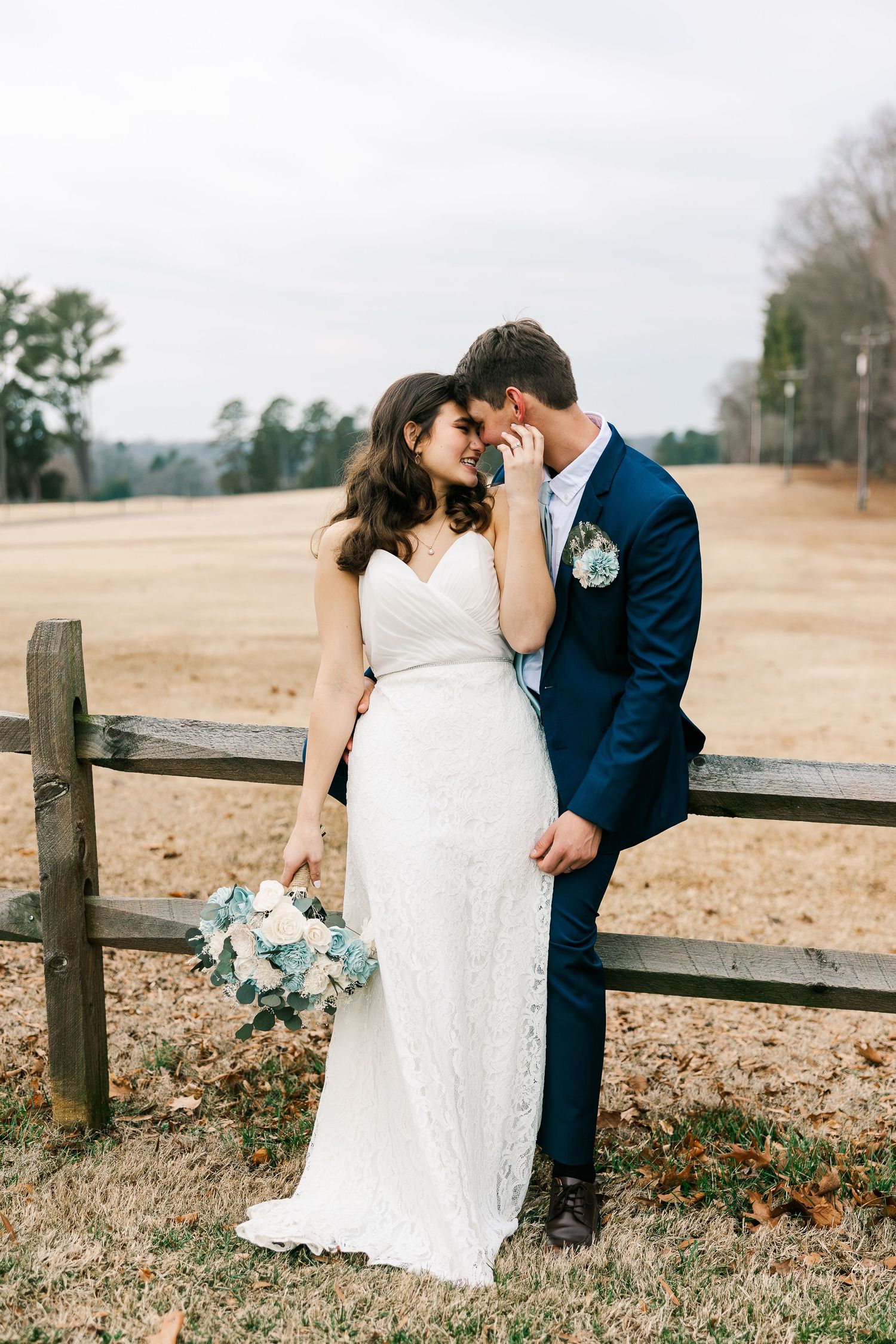 Bride in white lace gown and groom in navy suit share a romantic moment by a wooden fence outdoors.