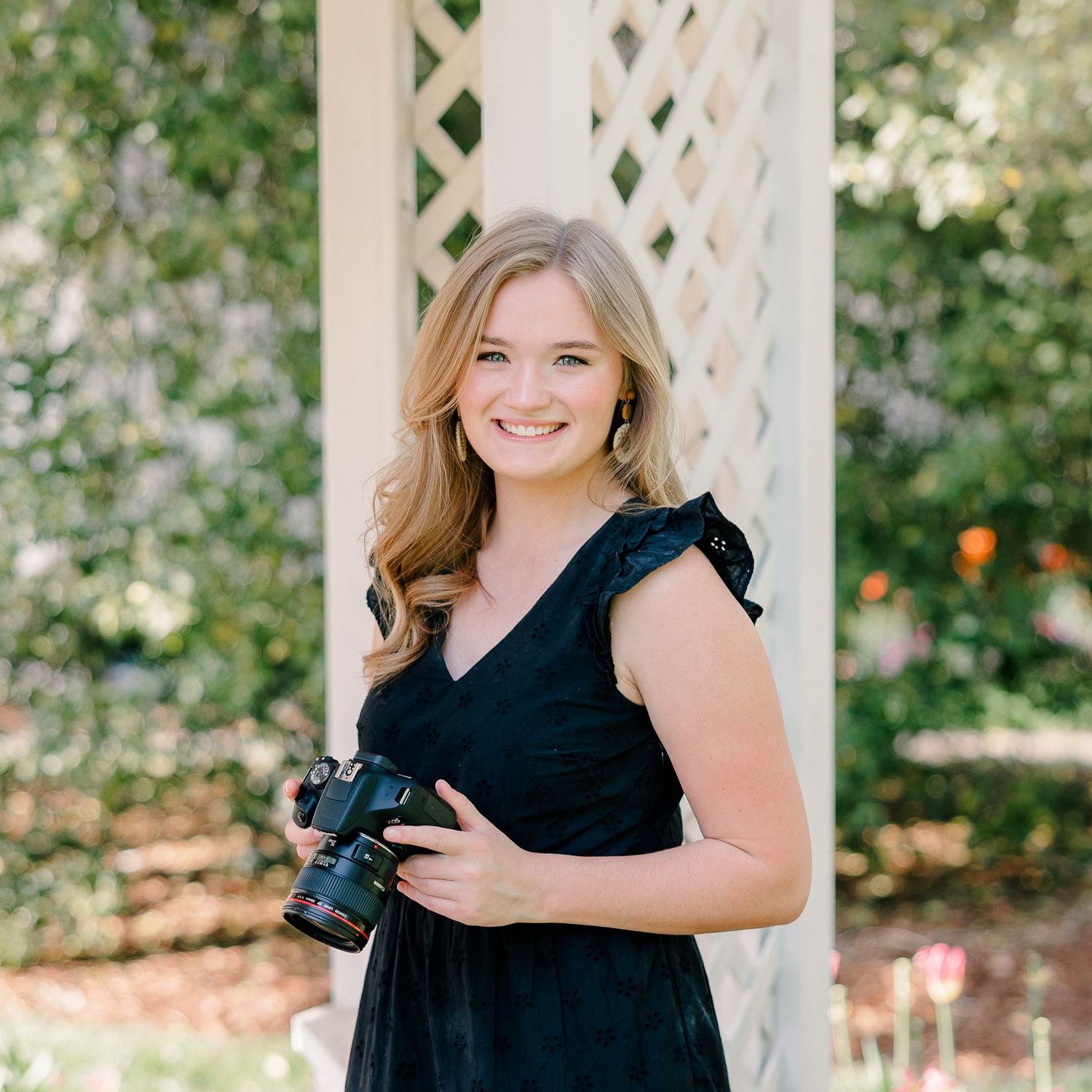Young blonde woman in black dress holding a camera, smiling outdoors near white lattice arbor surrounded by greenery.