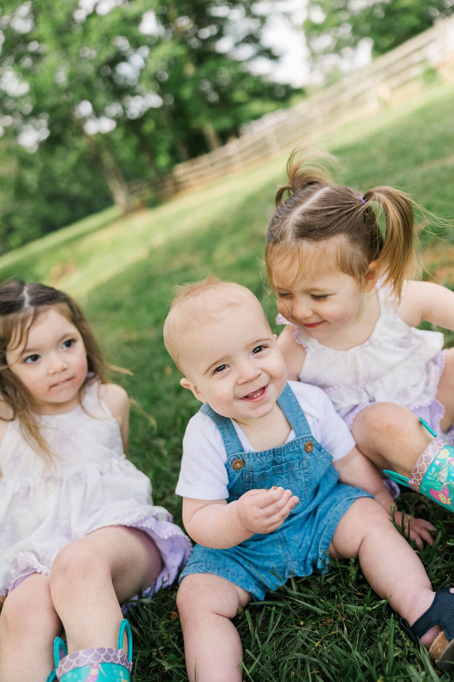 Three young children sitting together on green grass outdoors, smiling and interacting in a sunny park setting.