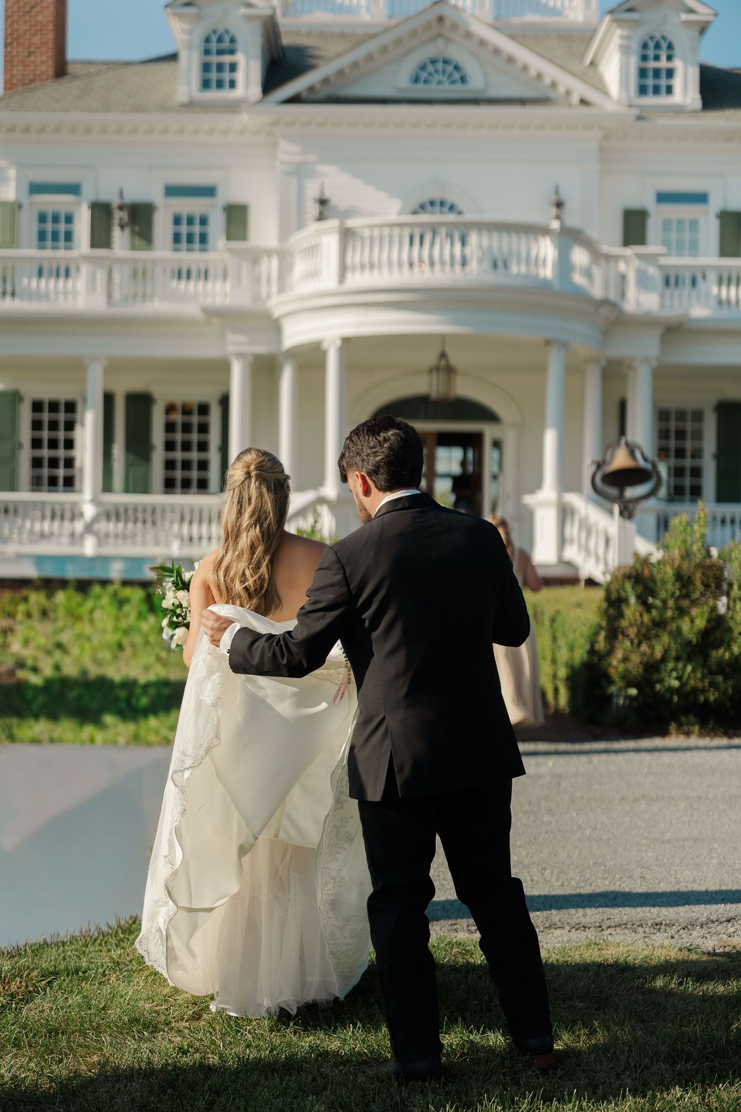 Bride in a flowing white gown and groom in a black suit walk arm in arm toward a grand white mansion with balconies and columns.