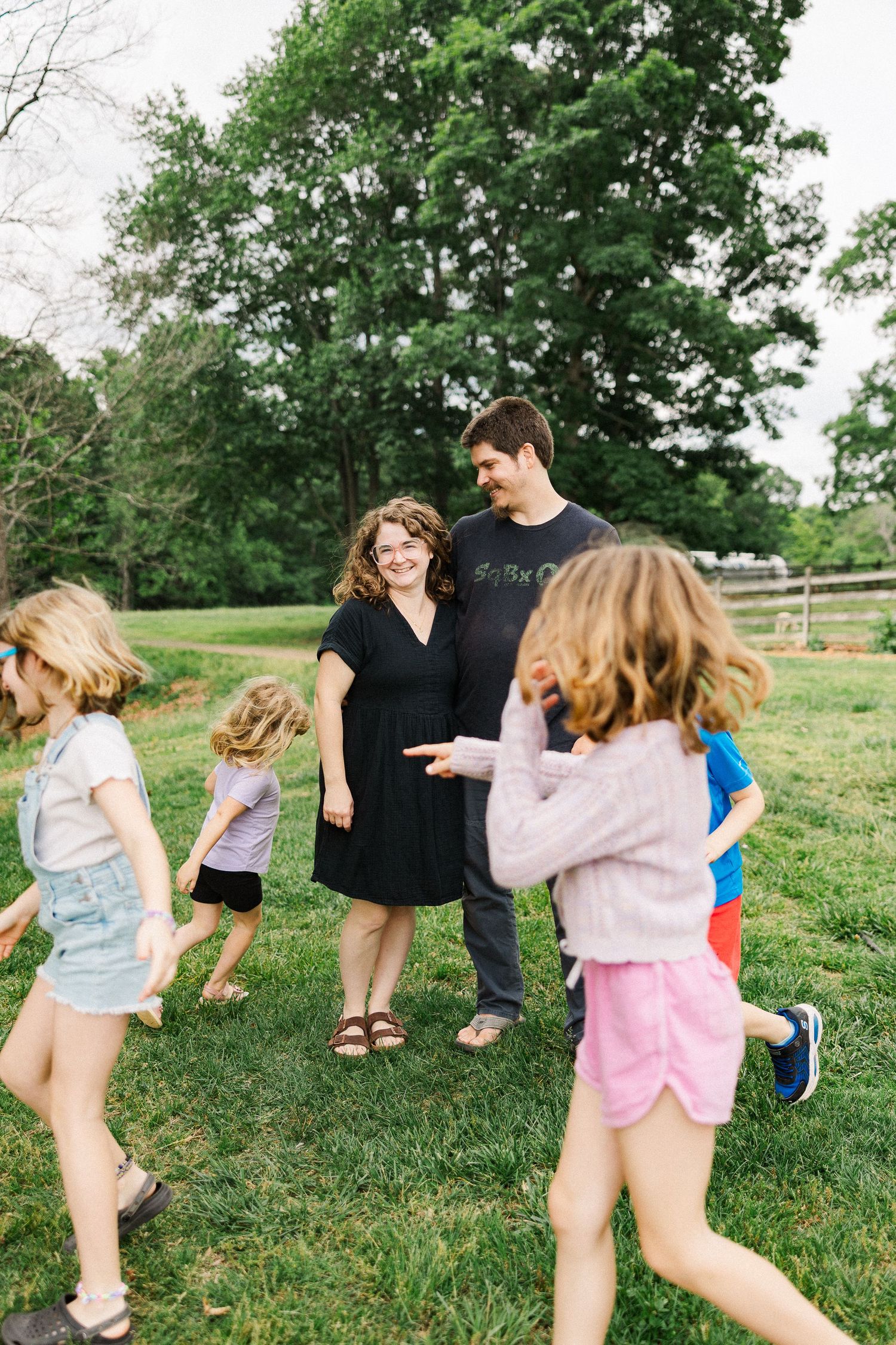 A family with children playing outdoors on green grass surrounded by tall trees on a sunny day.