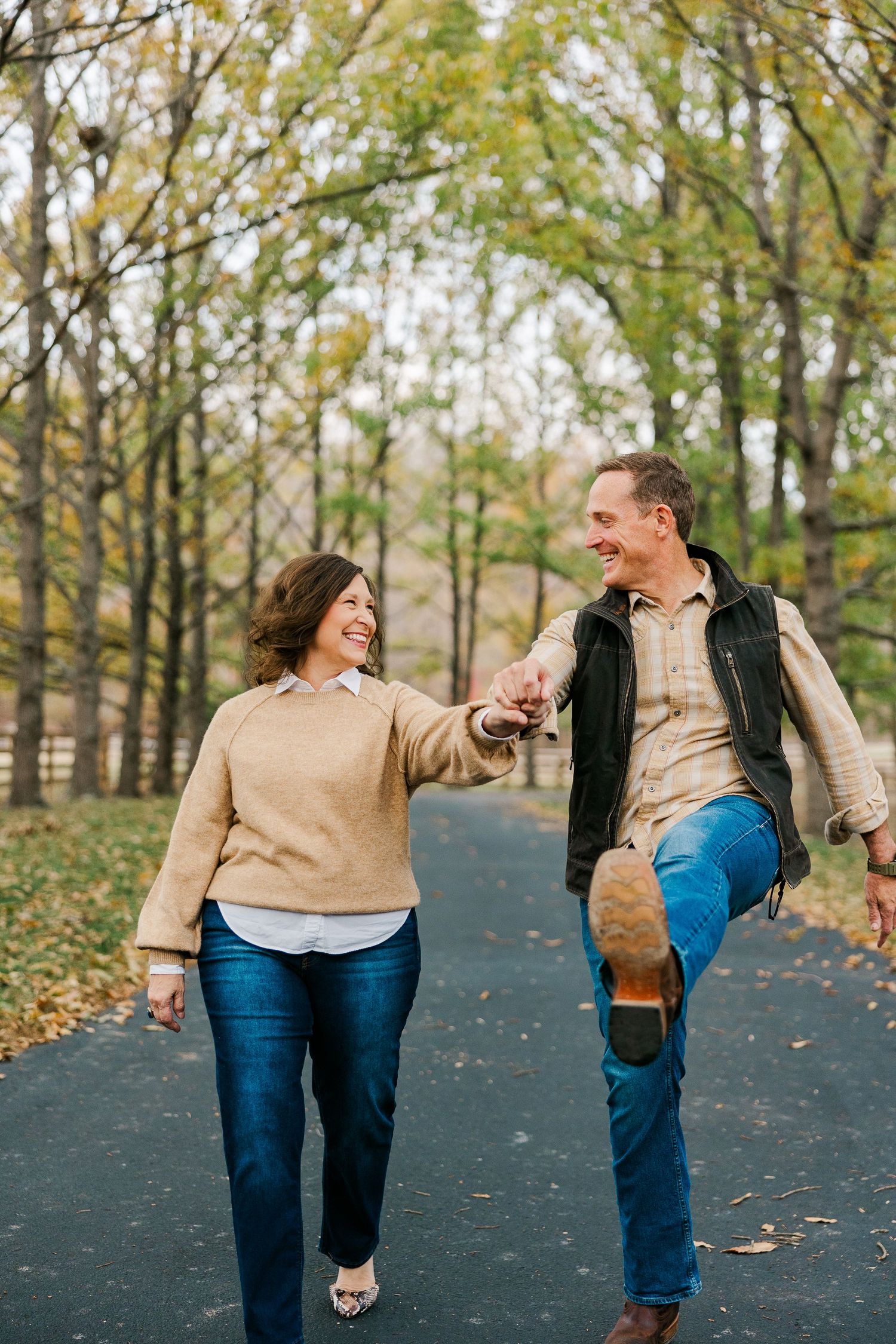 A joyful couple holds hands and laughs while walking on a tree-lined path during autumn.